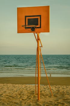 A solitary basketball hoop stands on a sandy beach against a tranquil sea backdrop.