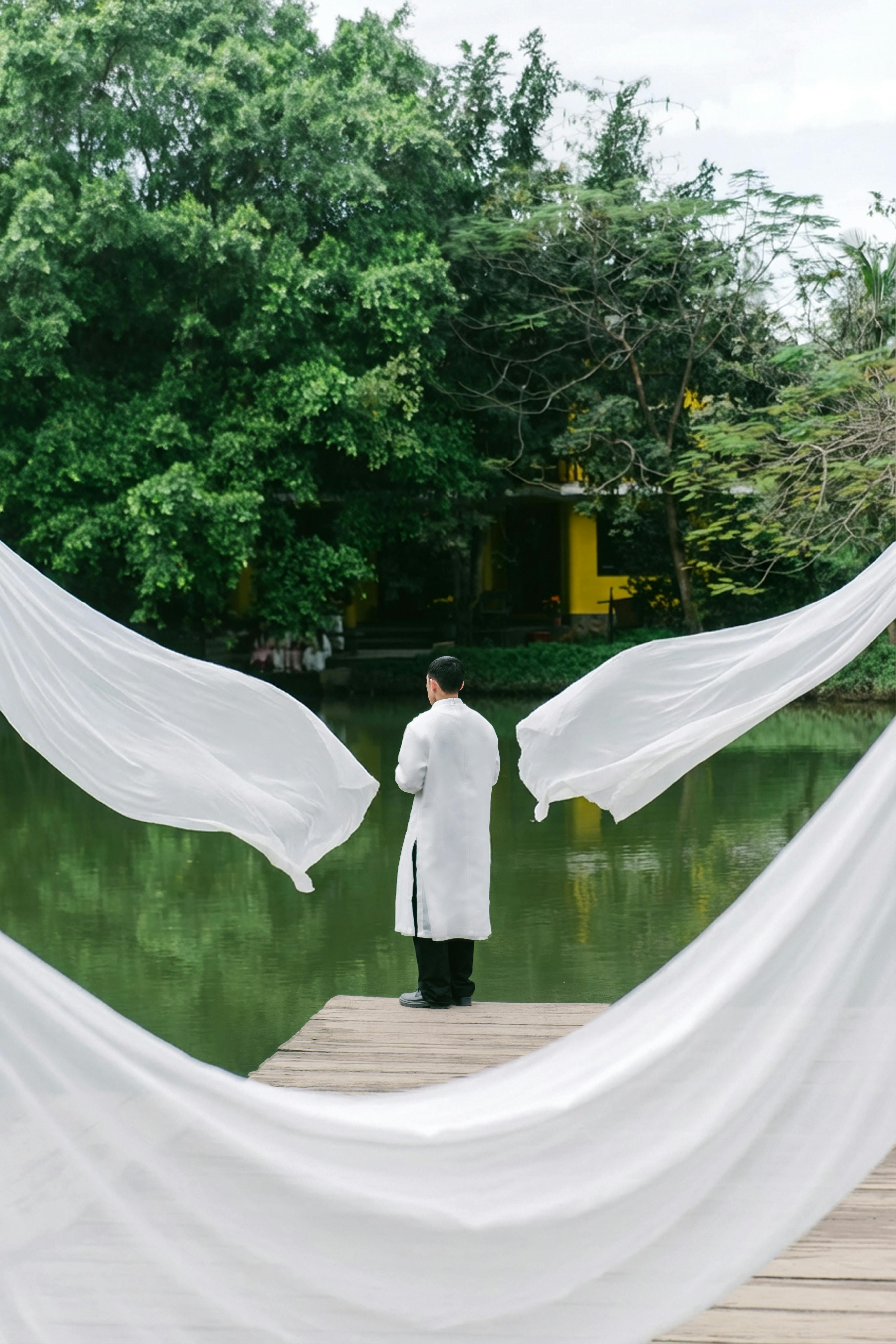 ingyenes Egy fehér köpenyes férfi áll egy mólón egy békés tó partján, lenge fehér anyag veszi körül. Stockfotó