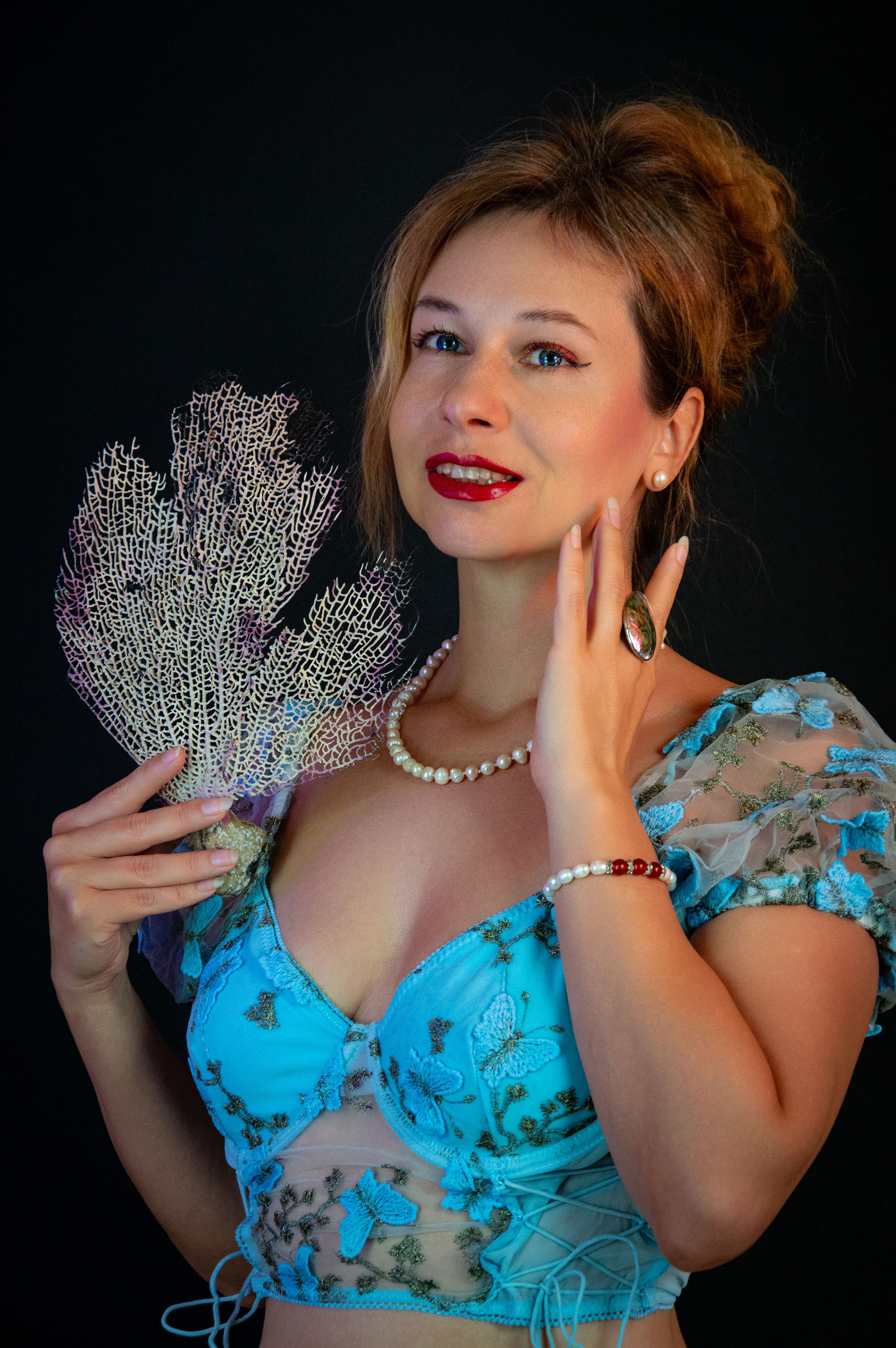Portrait of a woman in a blue embroidered top holding a coral fan.