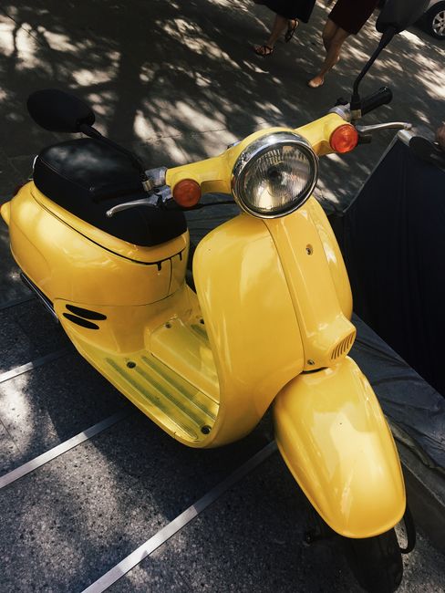 A bright yellow motor scooter parked outdoors under dappled sunlight on a city street.