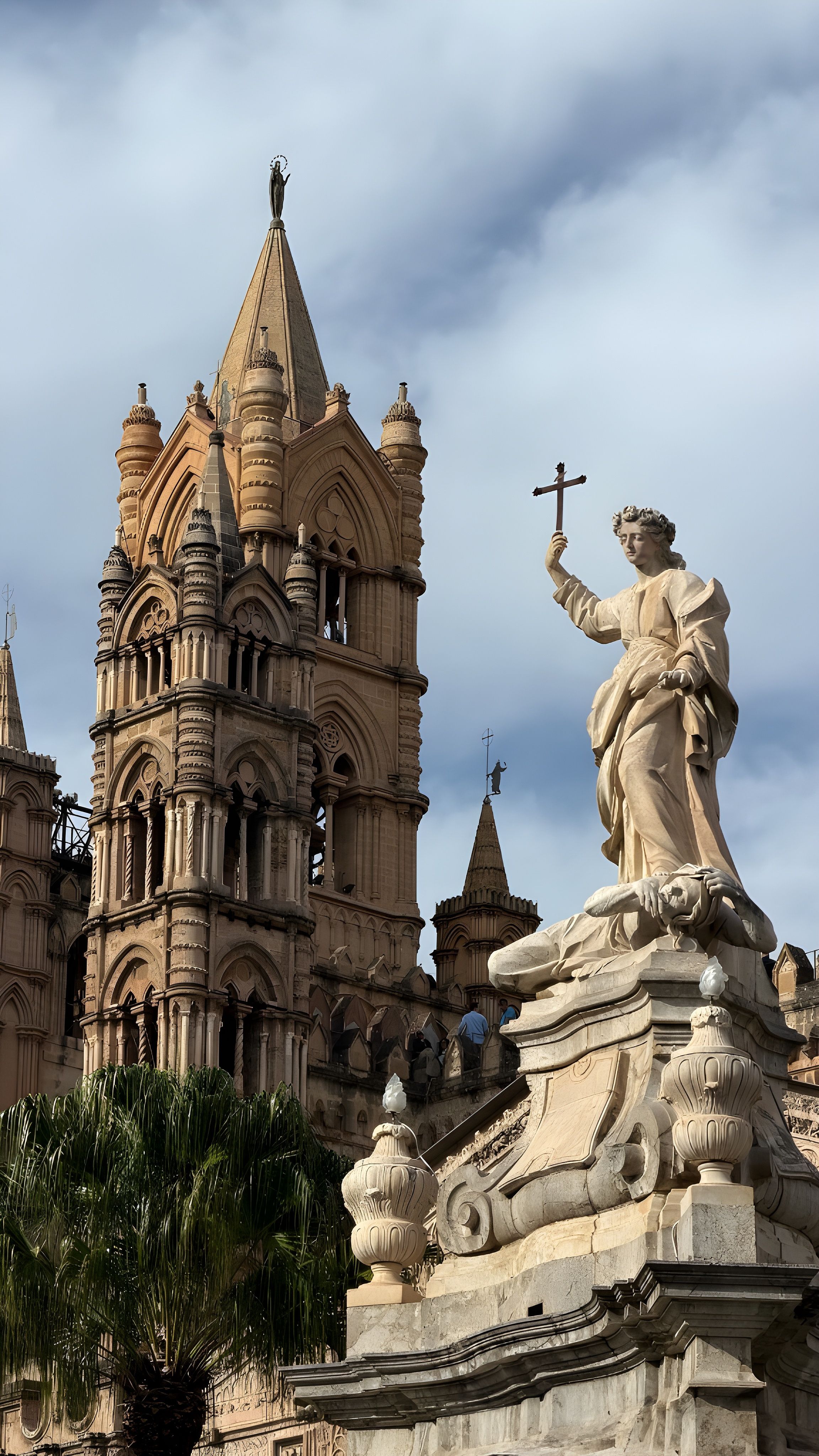 Historic Gothic cathedral tower and religious statue against a dramatic, cloudy sky.