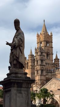 Statue and historic cathedral with Gothic architecture in Palermo under cloudy skies.