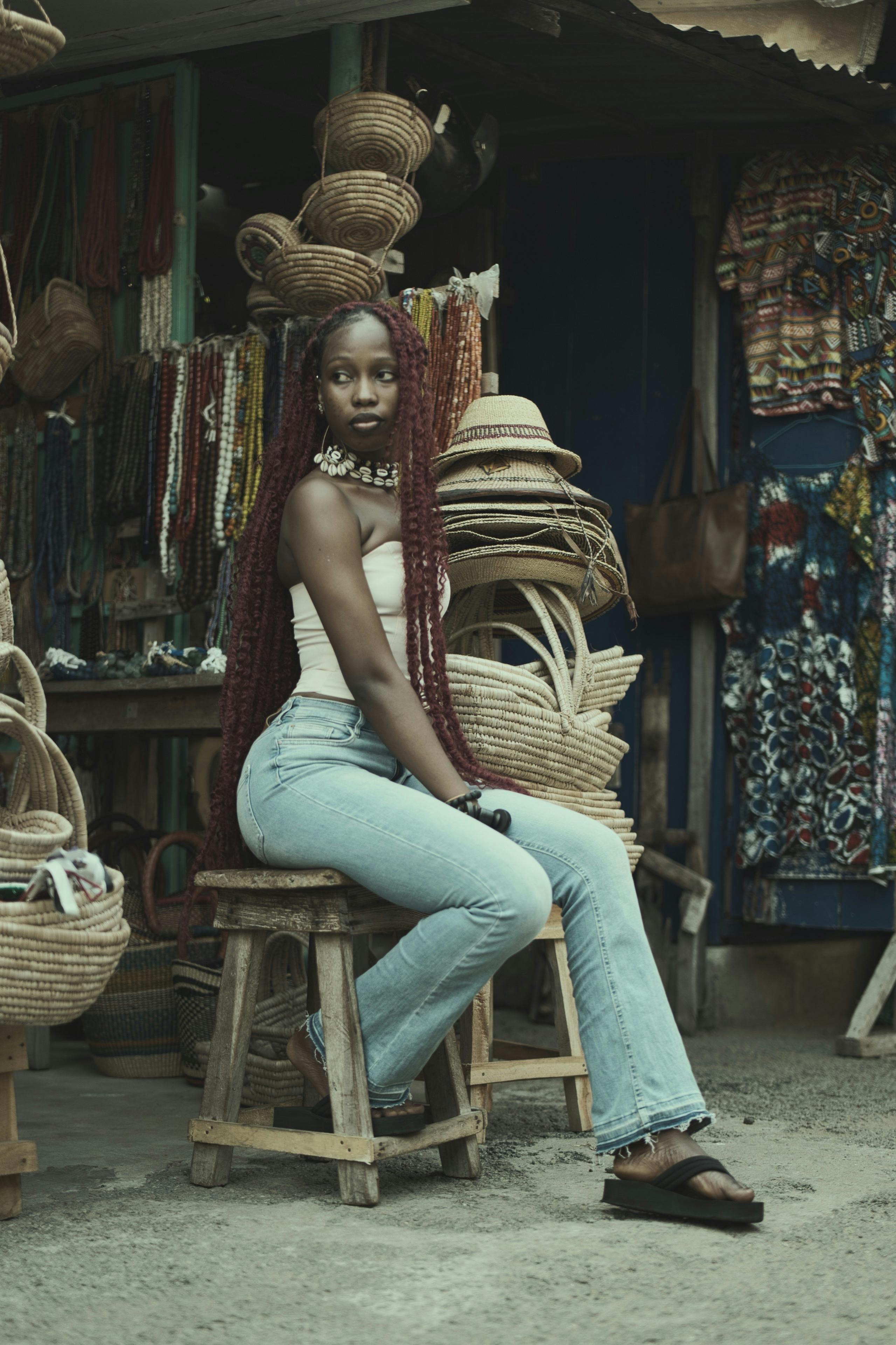 ฟรี หญิงสาวแต่งตัวมีสไตล์นั่งอยู่บนเก้าอี้เตี้ย ท่ามกลางสินค้าตลาดสีสันสดใส ในบรรยากาศกลางแจ้ง คลังภาพถ่าย