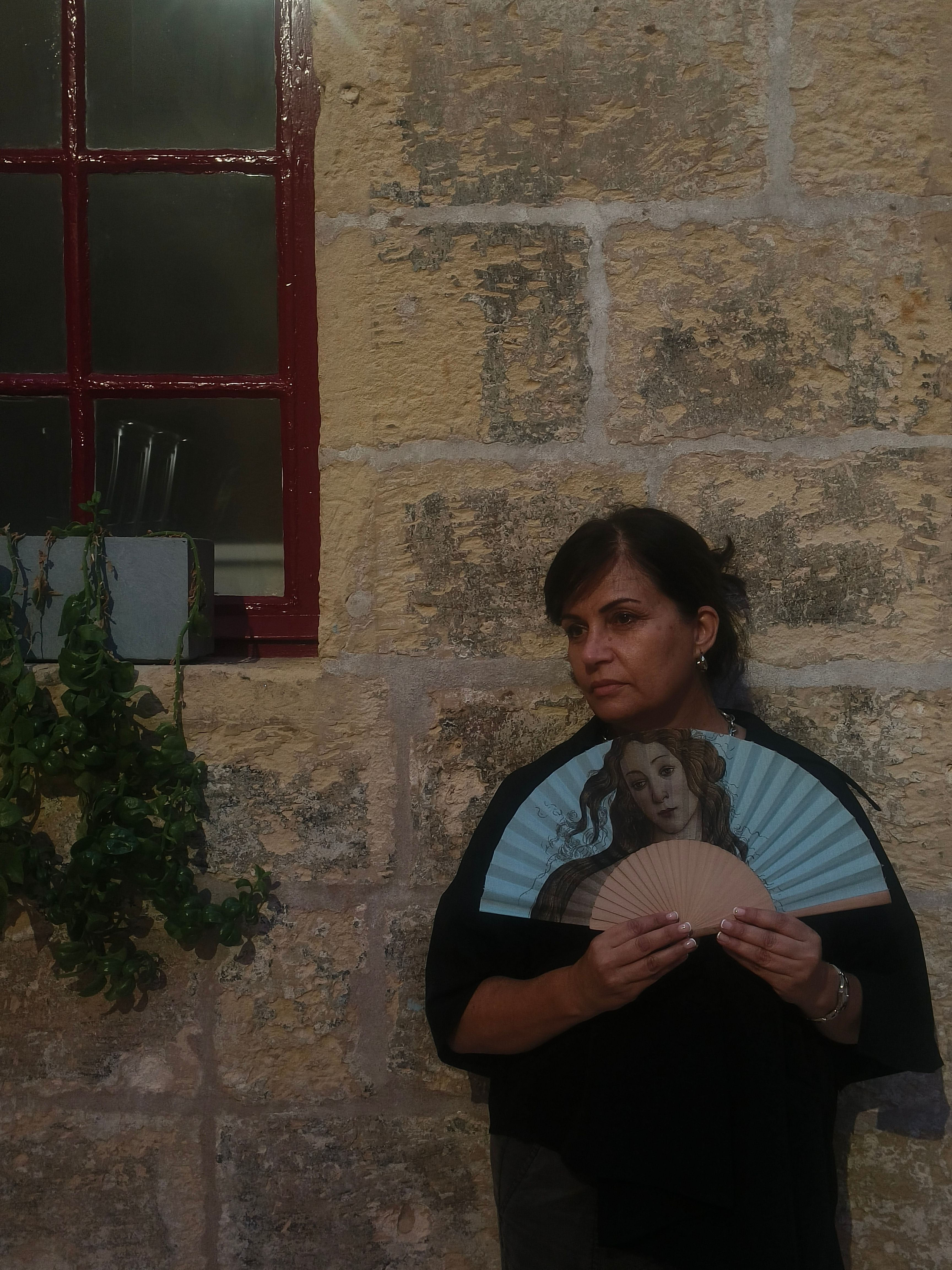 A woman holds a decorative fan against a historic stone wall beside a window.