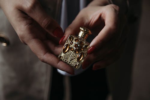 Crop faceless female with red manicure demonstrating small vintage golden perfume bottle decorated with natural stones and carved details