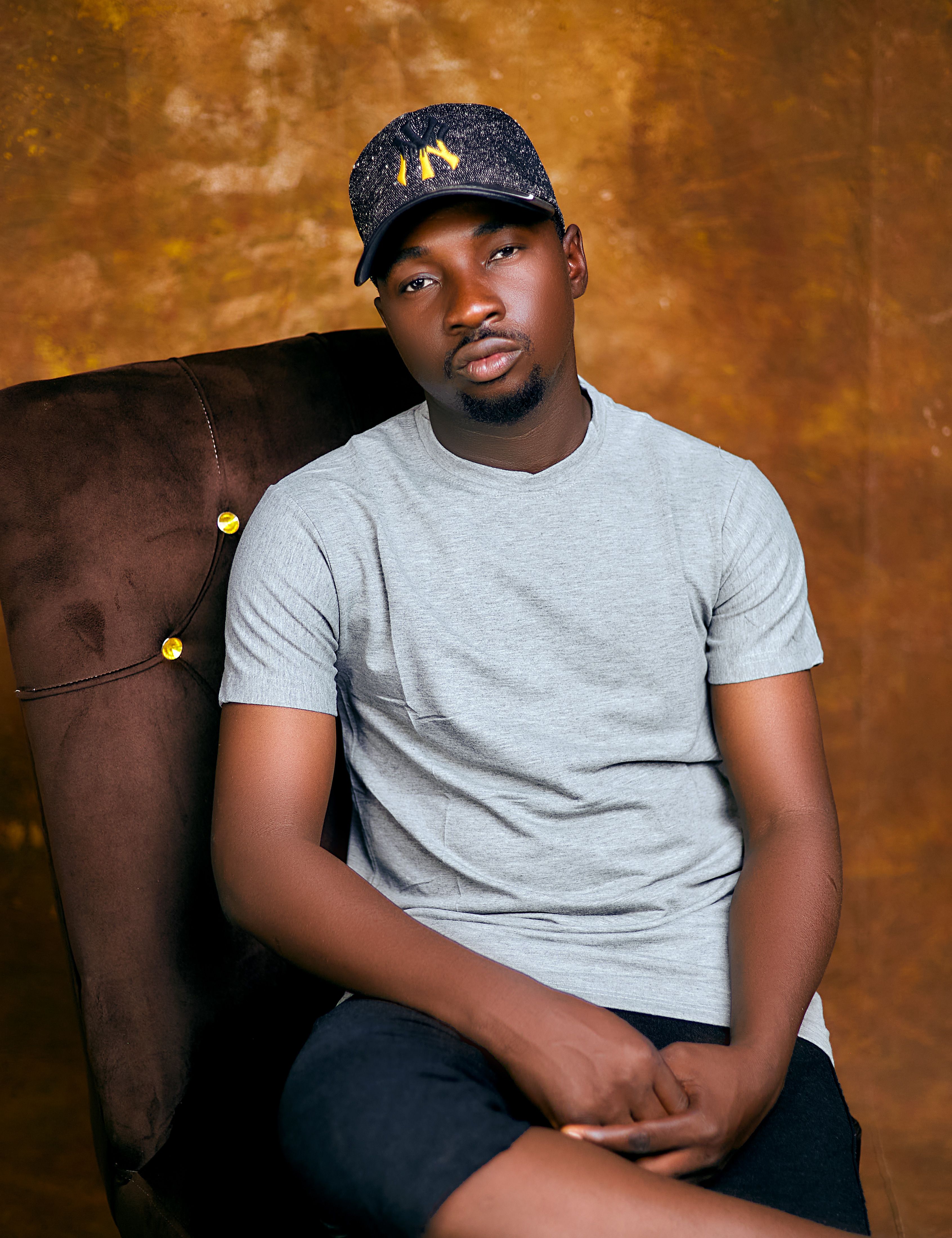 Young man in gray shirt and cap sitting on a brown chair against a warm backdrop.