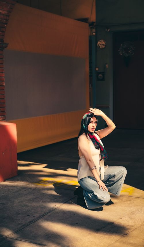 Free Fashionable woman with colorful scarf kneeling in urban setting, highlighting trendy style and sunlight contrast. Stock Photo