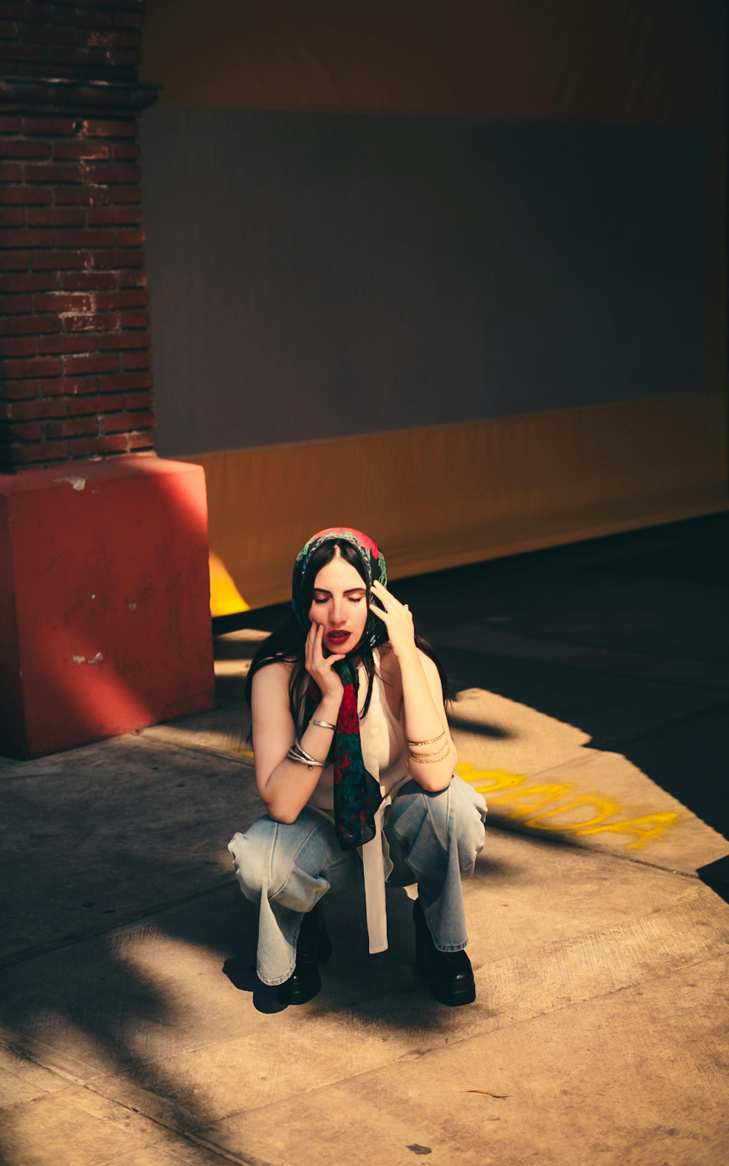 Bezpłatne Młoda kobieta siedzi zamyślona w nasłonecznionym miejskim otoczeniu, emanując swobodną, ​​artystyczną atmosferą. Zdjęcie z galerii