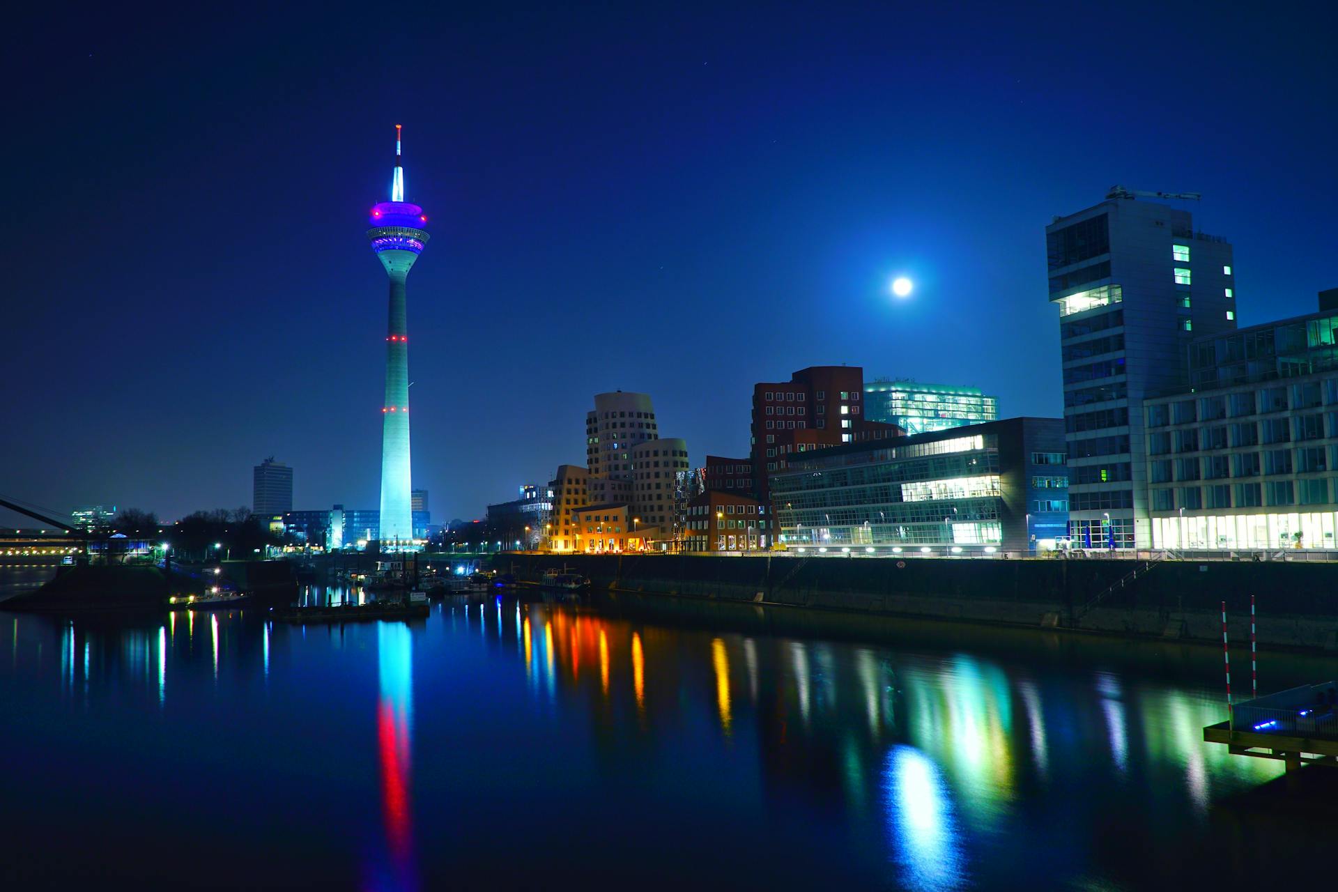 Scenic nighttime view of the Düsseldorf skyline featuring the illuminated Rhine Tower reflected in the river.