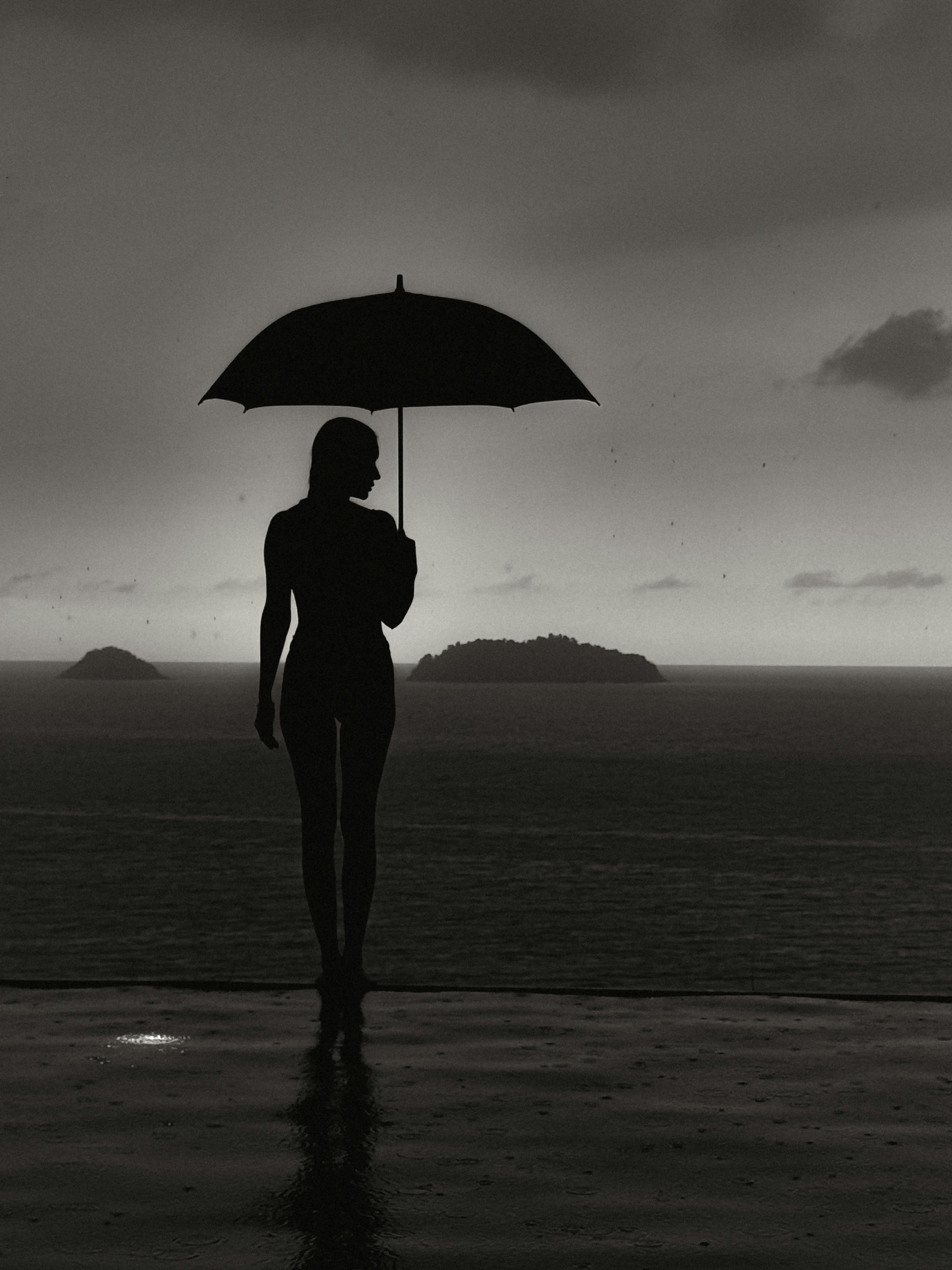 Gratis Elegante silhouette di una donna che tiene un ombrello in riva al mare, con isole in lontananza. Foto a disposizione