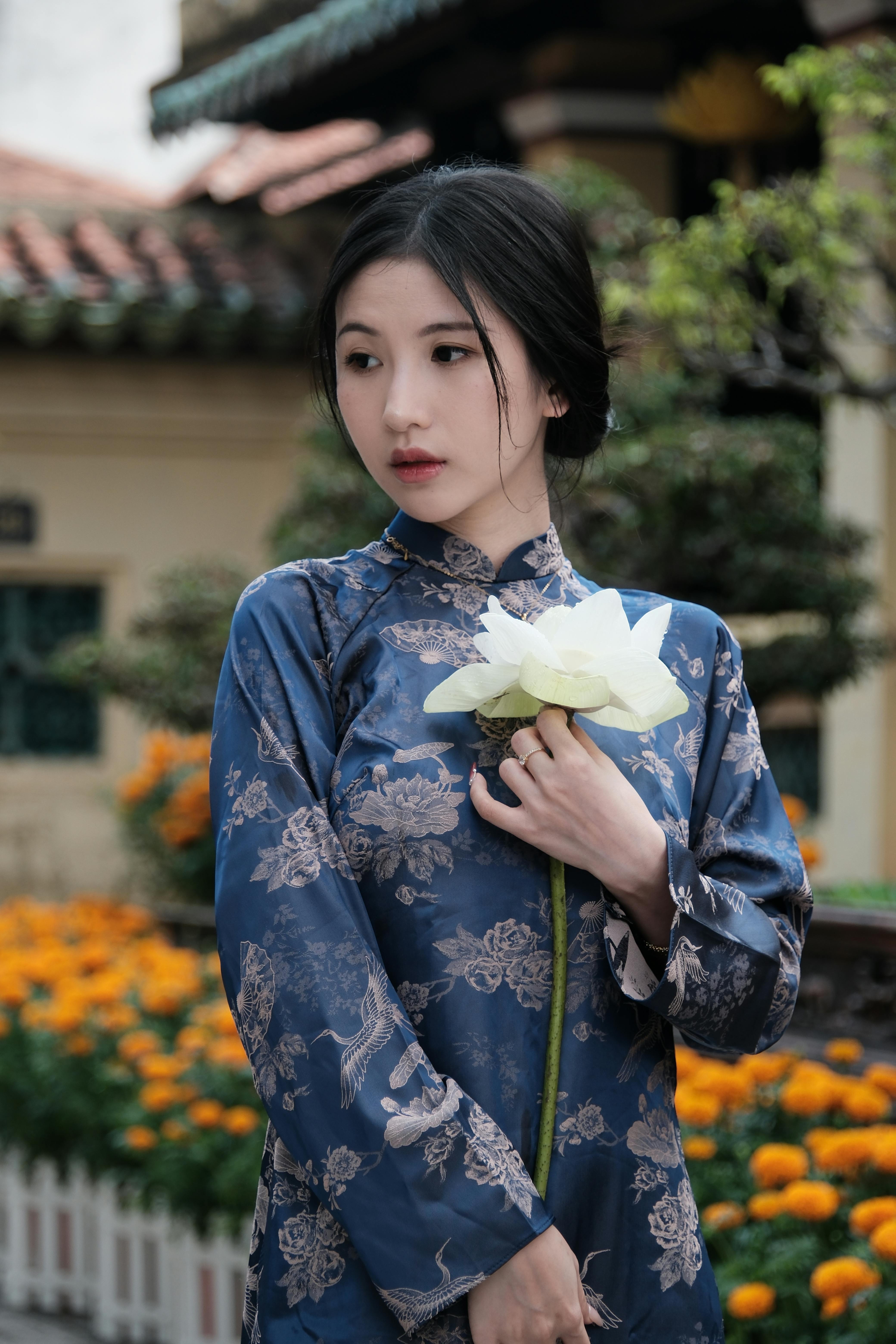 Gratis Una donna con un abito tradizionale blu tiene in mano un loto bianco, circondata da fiori dai colori vivaci all'aperto. Foto a disposizione