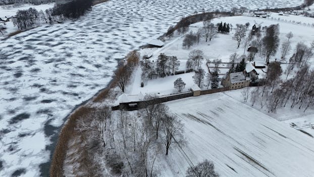 Aerial view of a snowy landscape featuring a frozen river and rural architecture in Poland.