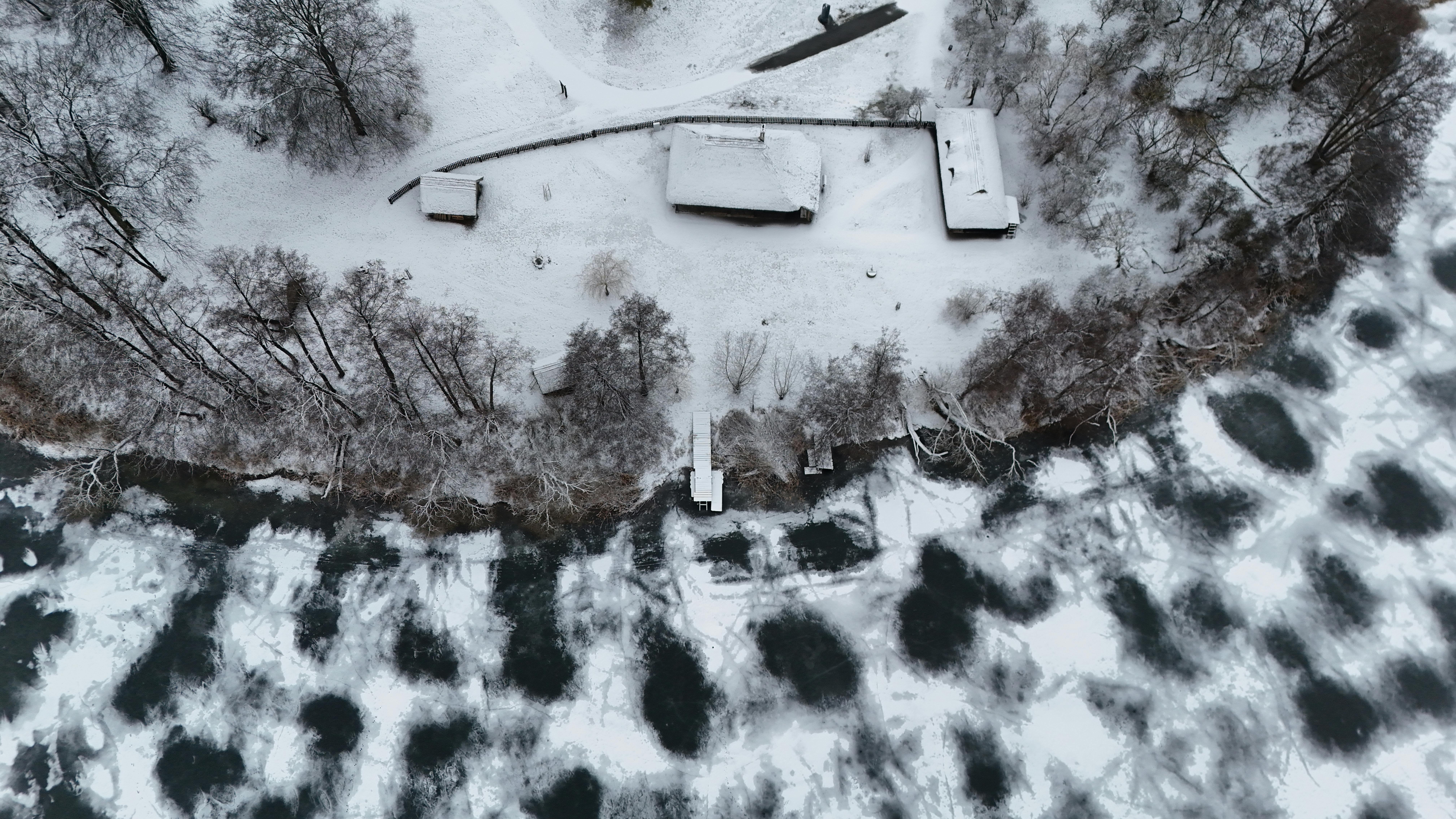 bezplatná Dron zachytil zasněženou chatu u zamrzlé řeky, obklopenou stromy v poklidné zimní krajině. Základová fotografie