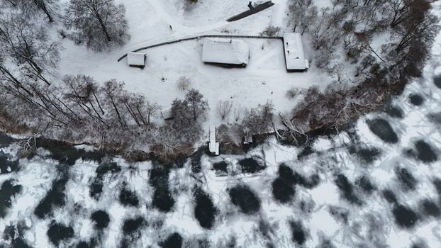 A drone captures a snowy cabin by a frozen river, surrounded by trees in winter's serene landscape.