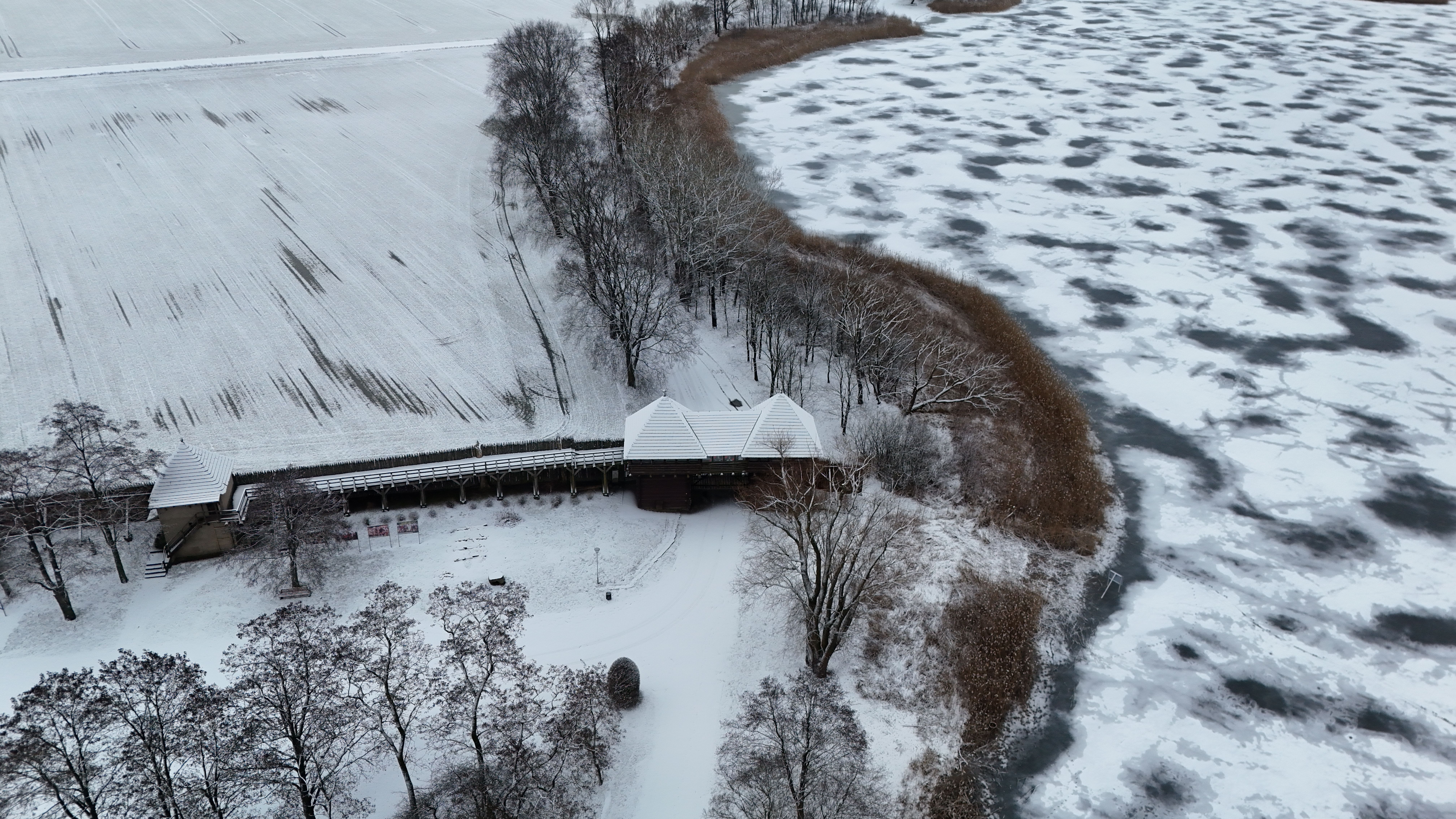 A winter aerial view of a snow-covered lakeside landscape with trees and a wooden structure. — Trójmiasto