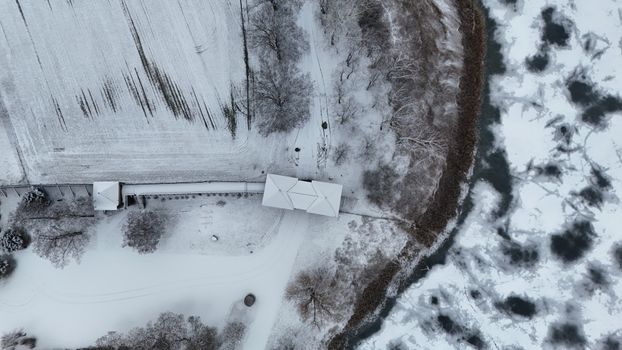 Aerial shot of a snowy rural area with a frozen river, trees, and small building.