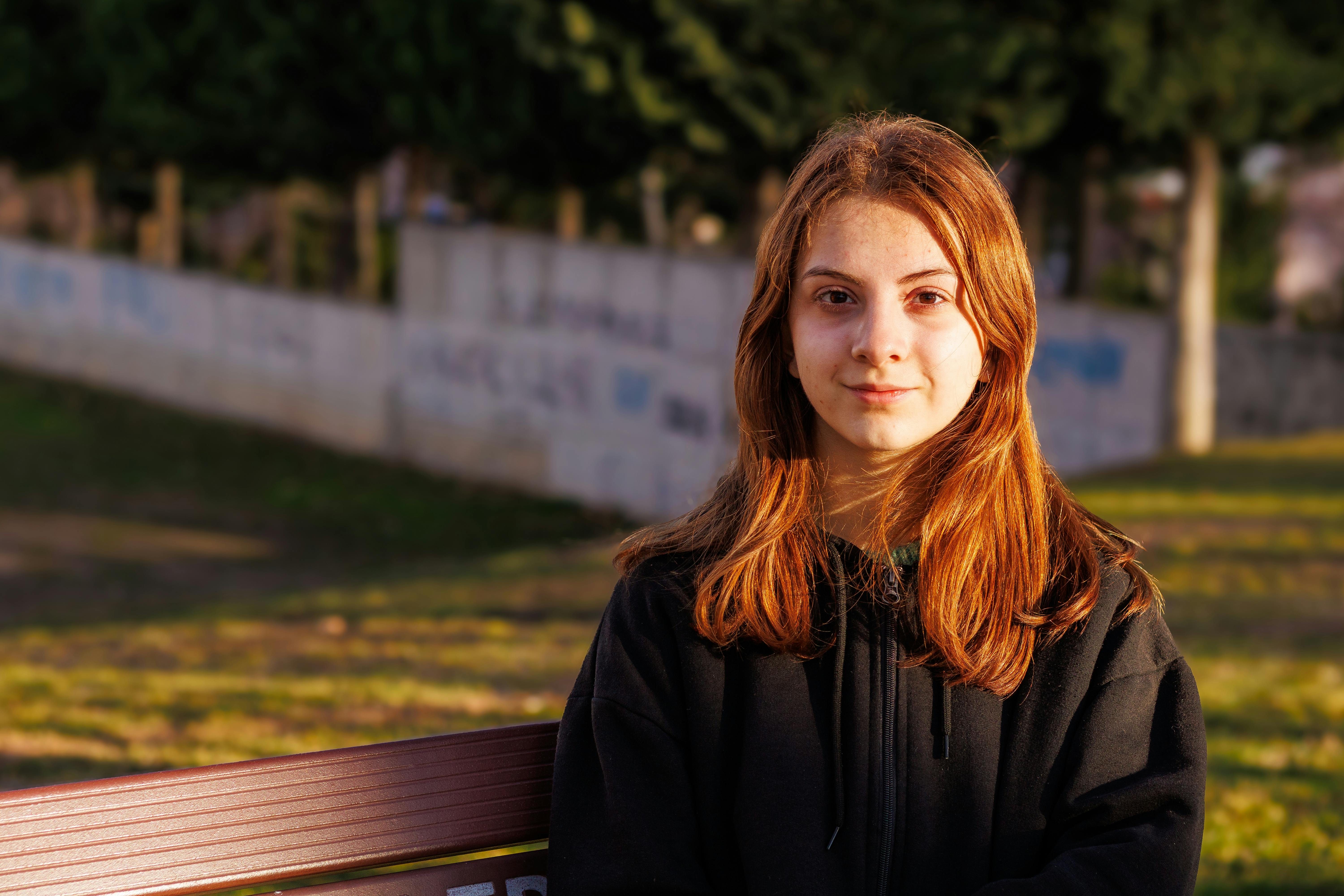 ฟรี หญิงสาวผมแดงนั่งอยู่บนม้านั่งในสวนสาธารณะที่มีแสงแดดส่องถึงในเมืองคอนยา ประเทศตุรกี คลังภาพถ่าย