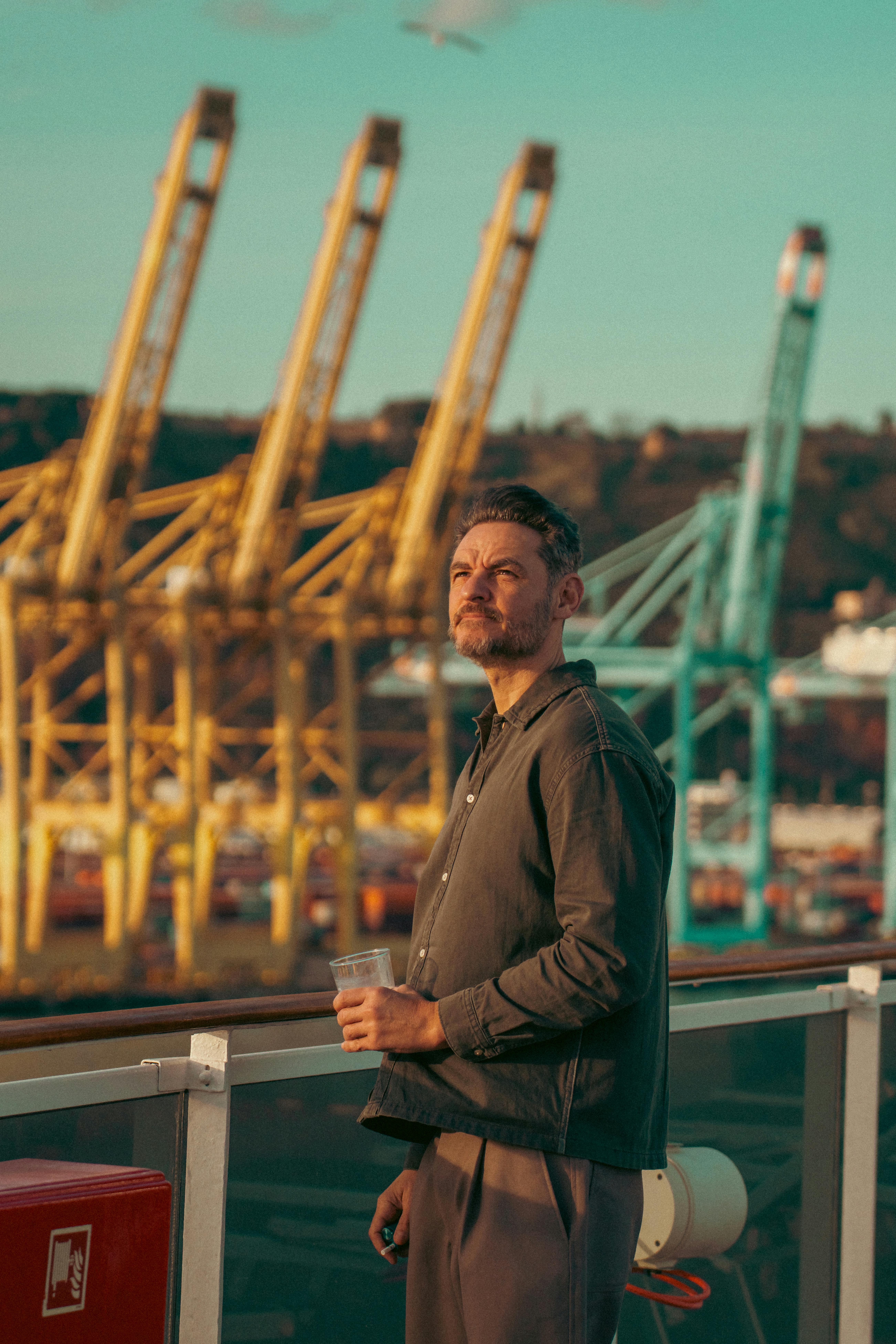 Free Man holding cup on balcony, overlooking colorful cranes at Spanish harbor. Stock Photo