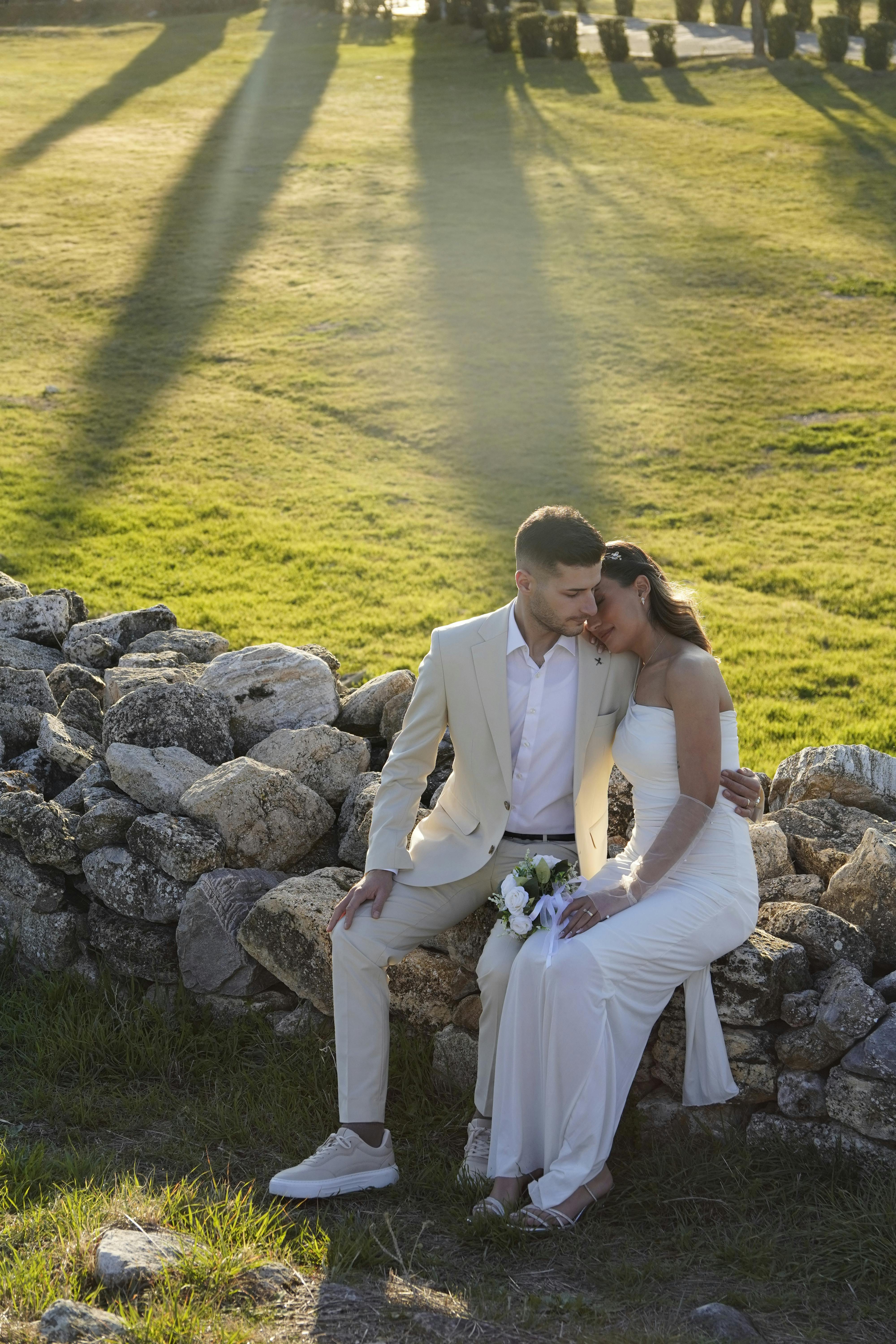 A couple in formal attire sharing a tender moment on a stone wall during sunset.
