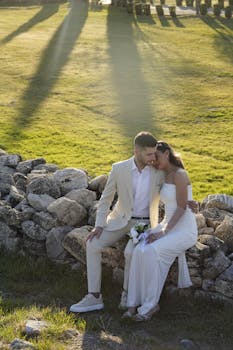 A couple in formal attire sharing a tender moment on a stone wall during sunset.