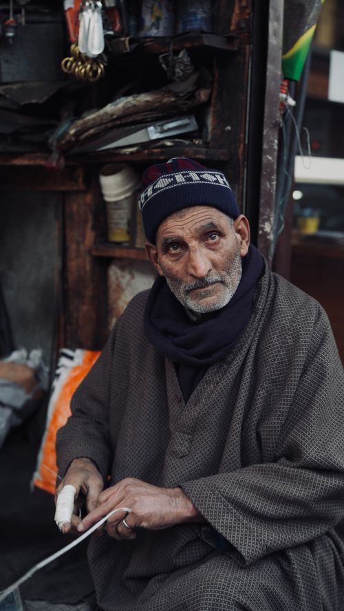 Free An elderly man wearing traditional Kashmiri attire sits in a market stall, showcasing local culture. Stock Photo