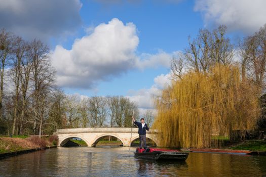 A person punts a boat under a stone bridge surrounded by trees on a sunny day.