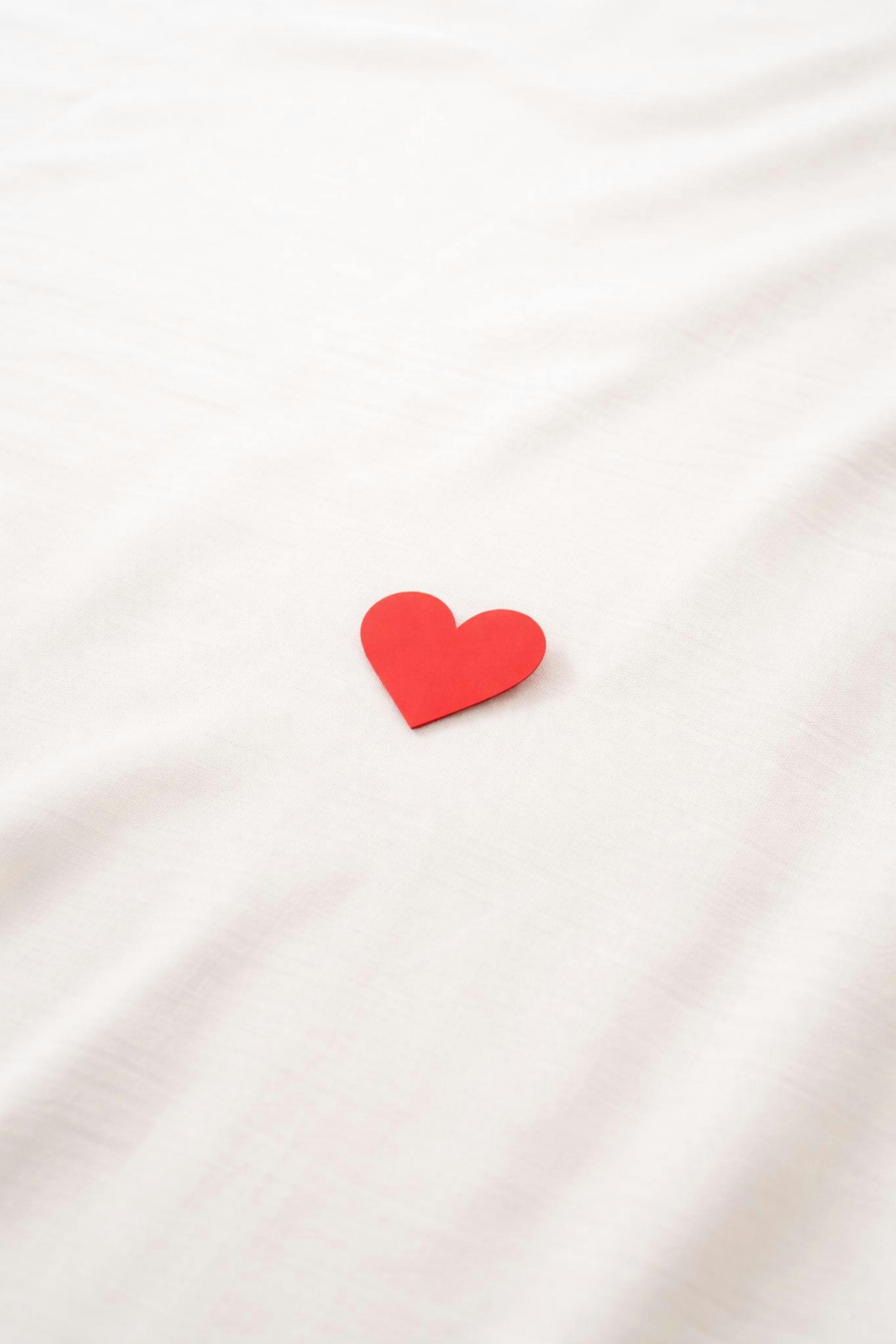 Ücretsiz Yumuşak beyaz bir kumaşın üzerinde duran minimalist kırmızı kağıt kalp, sevgiyi ve sadeliği simgeliyor. Stok Fotoğraflar