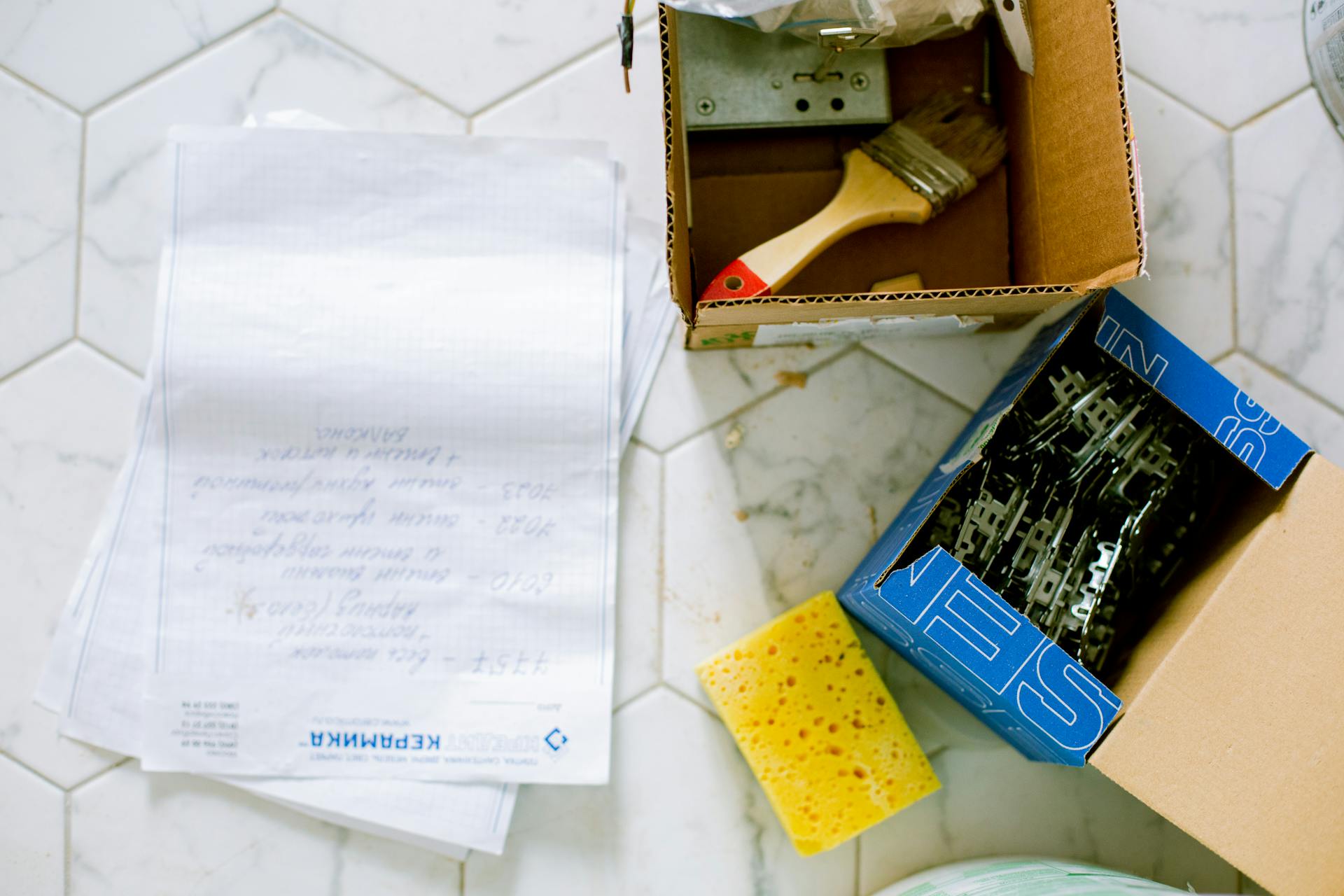 Flat lay of home renovation tools including paintbrush, sponge, and hardware on tiled floor.