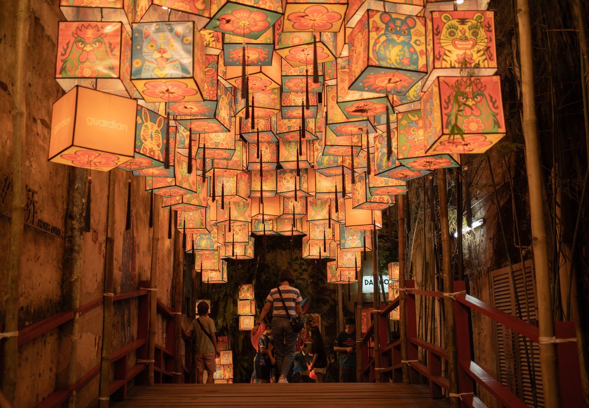 Dozens of painted paper box lanterns strung overhead down a narrow alley at dusk, people walking beneath them toward a lit doorway, the air carrying incense and someone's dinner frying two doors down.