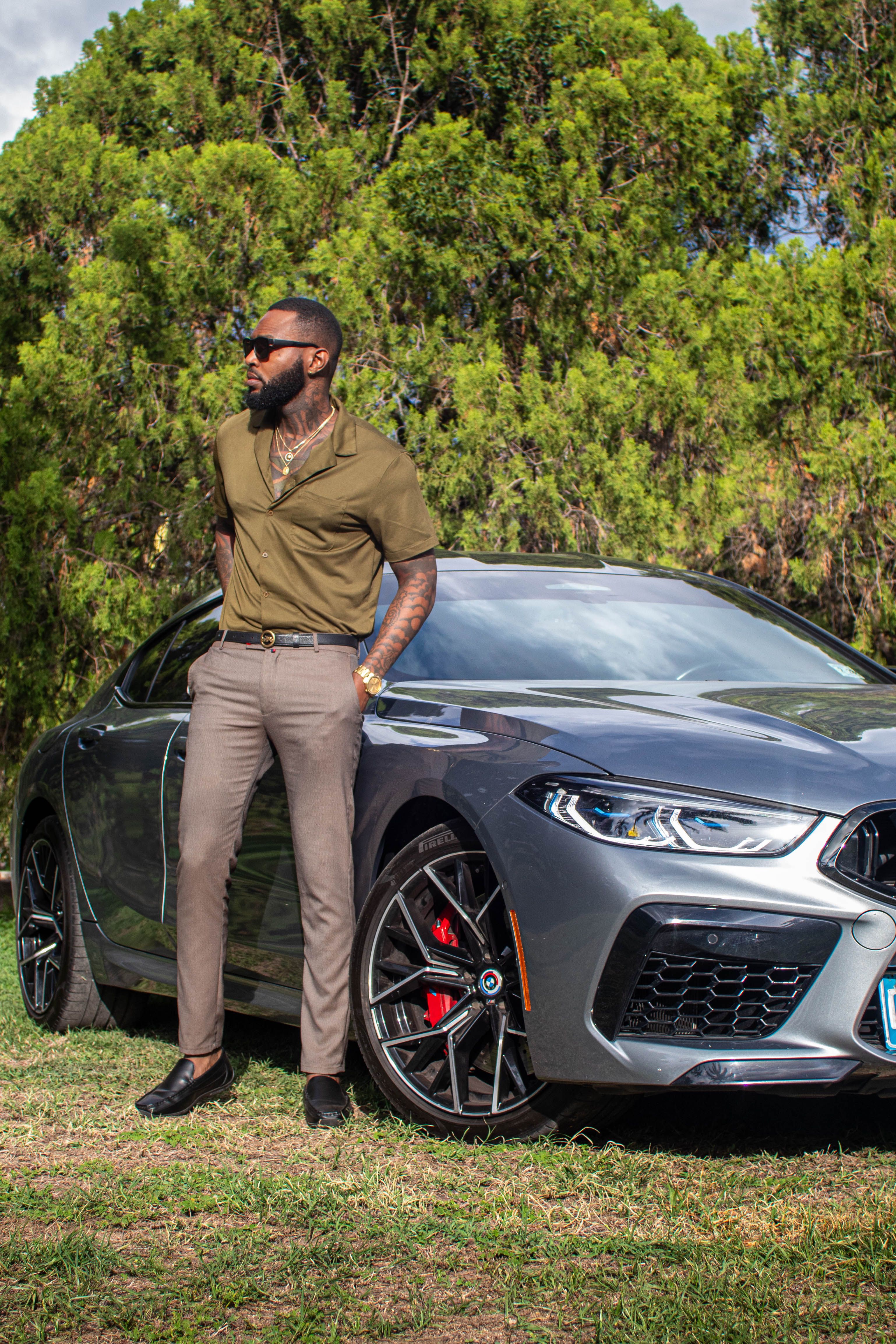 Fashionable man with beard and tattoos leans on a sleek luxury car outdoors.