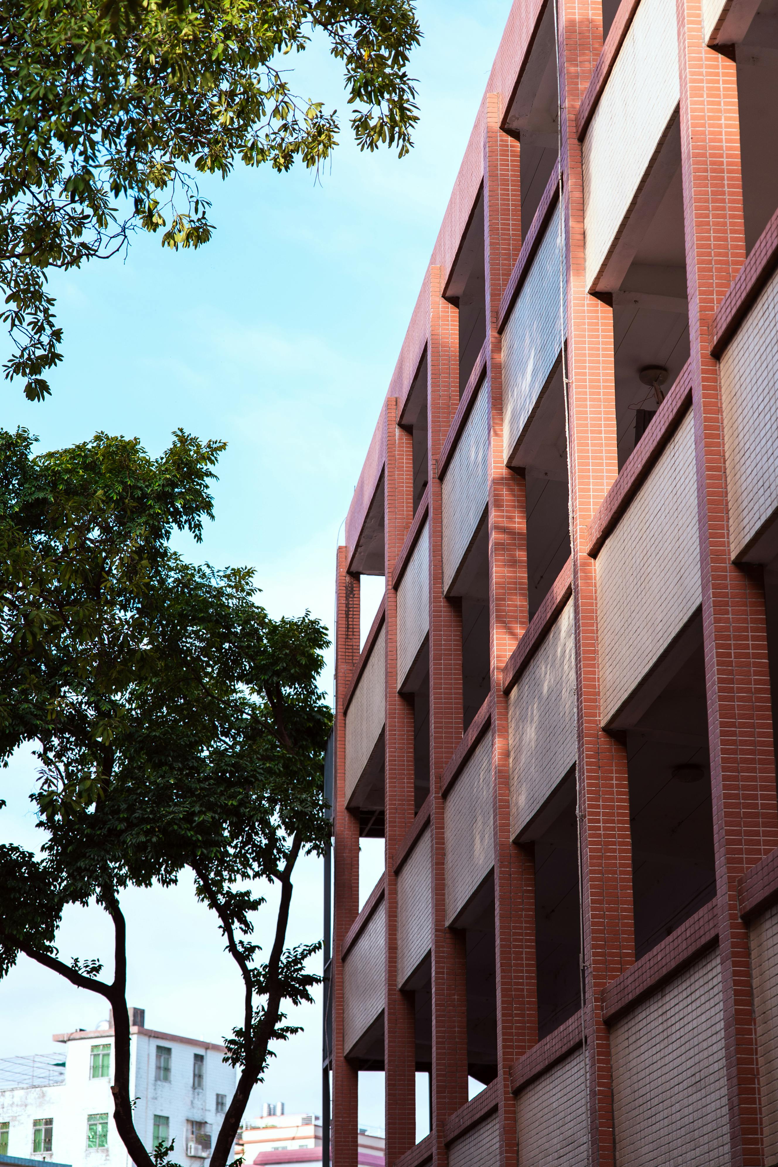 Gratis En moderne murstensbygning med åbne korridorer mod en klar blå himmel med træer i forgrunden. Lagerfoto