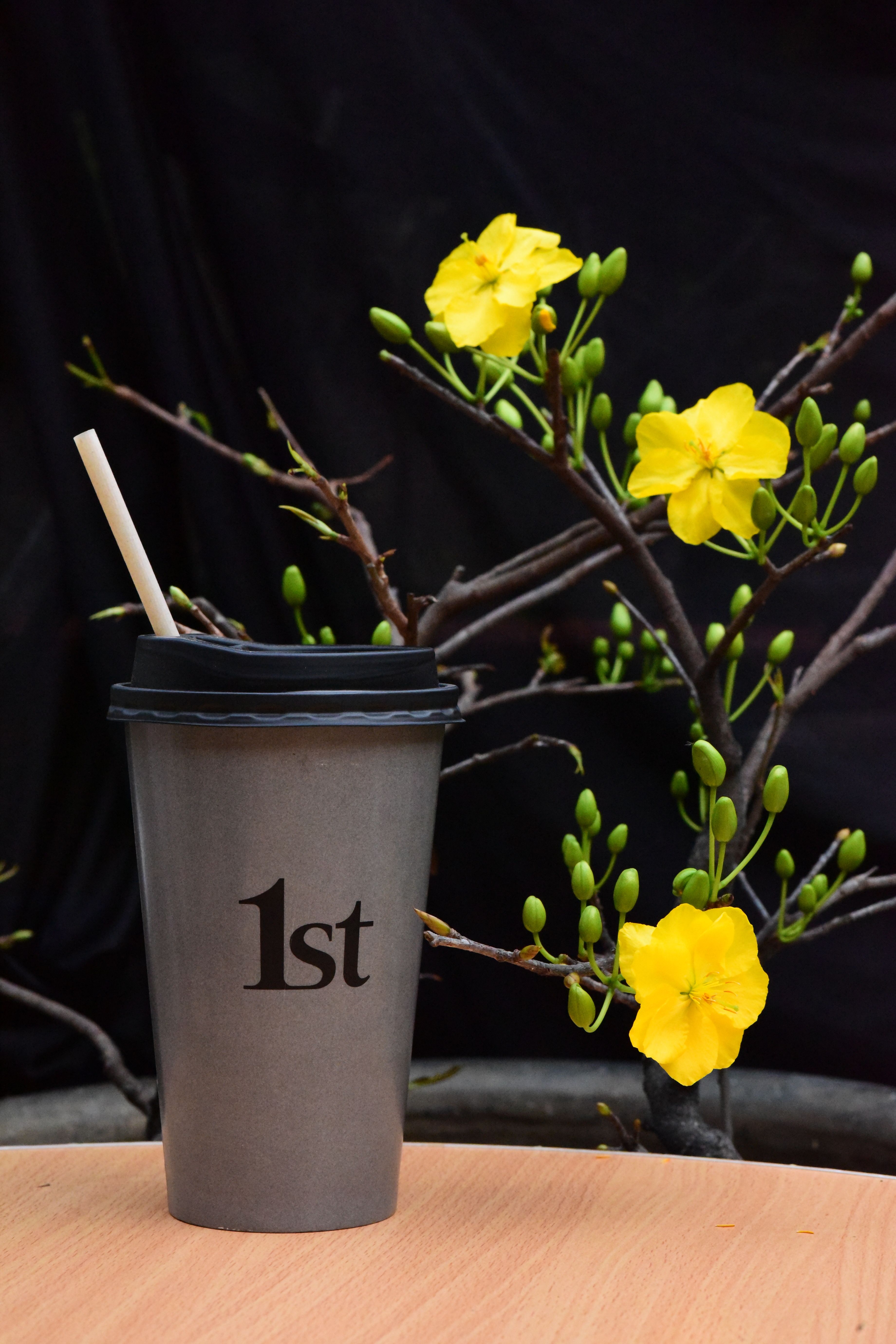 Black coffee cup with straw alongside bright yellow flowers against a dark background.