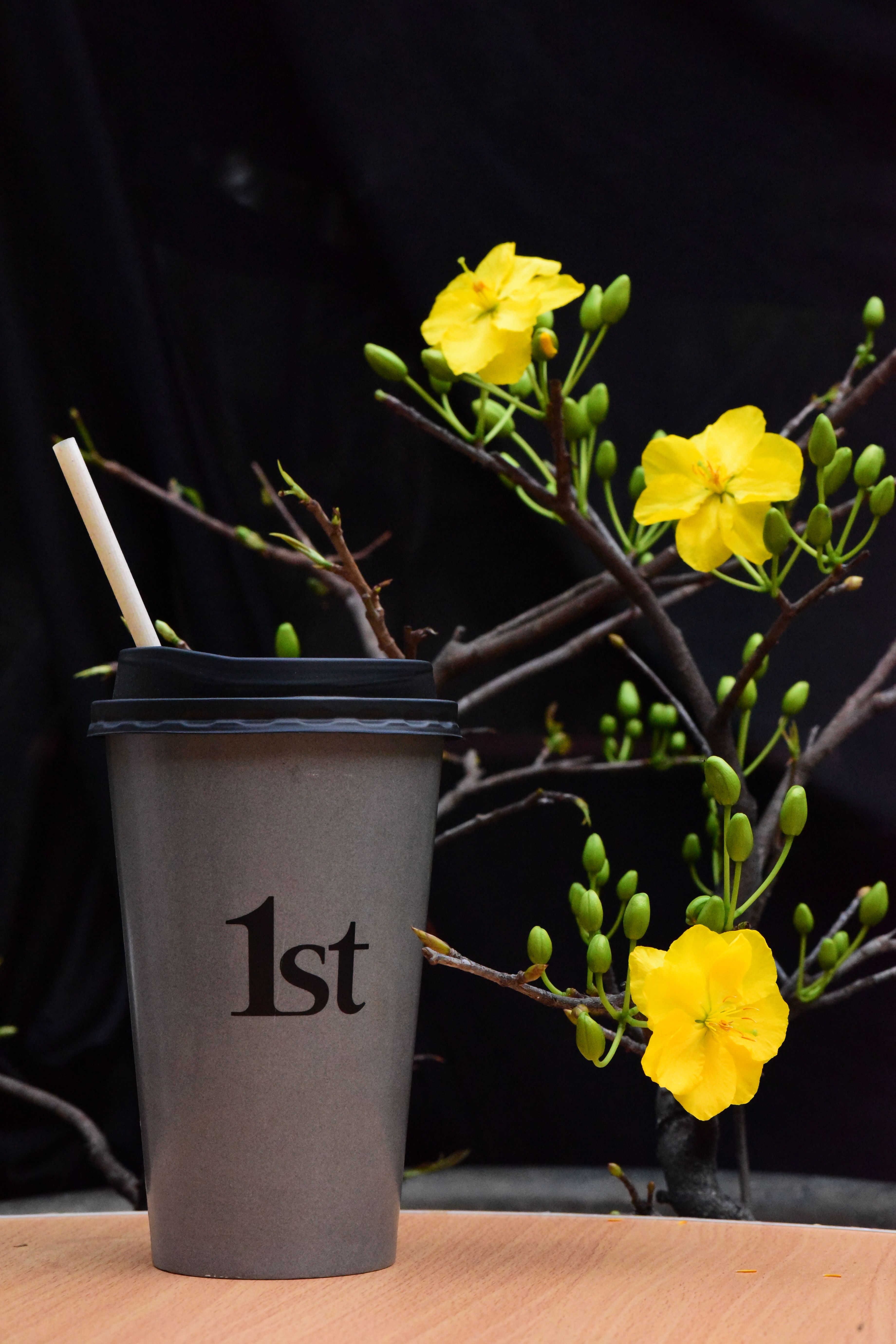 A gray coffee cup labeled '1st' stands beside vibrant yellow flowers on a black background.