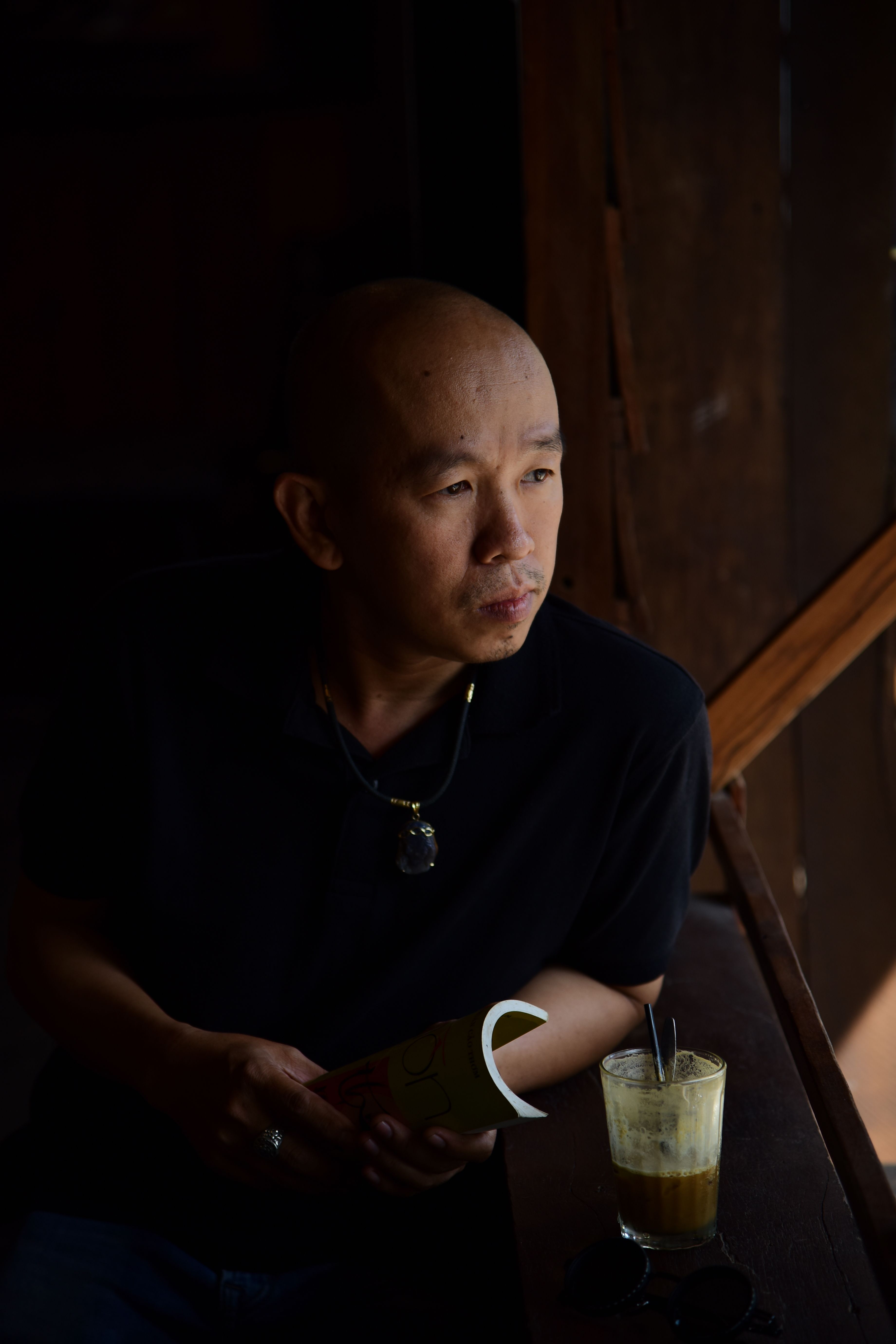 A man gazes out a window with a book and coffee in a dimly lit room.