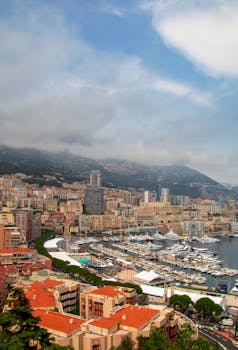 Aerial view of Monaco harbor with yachts and city buildings under a cloudy sky.