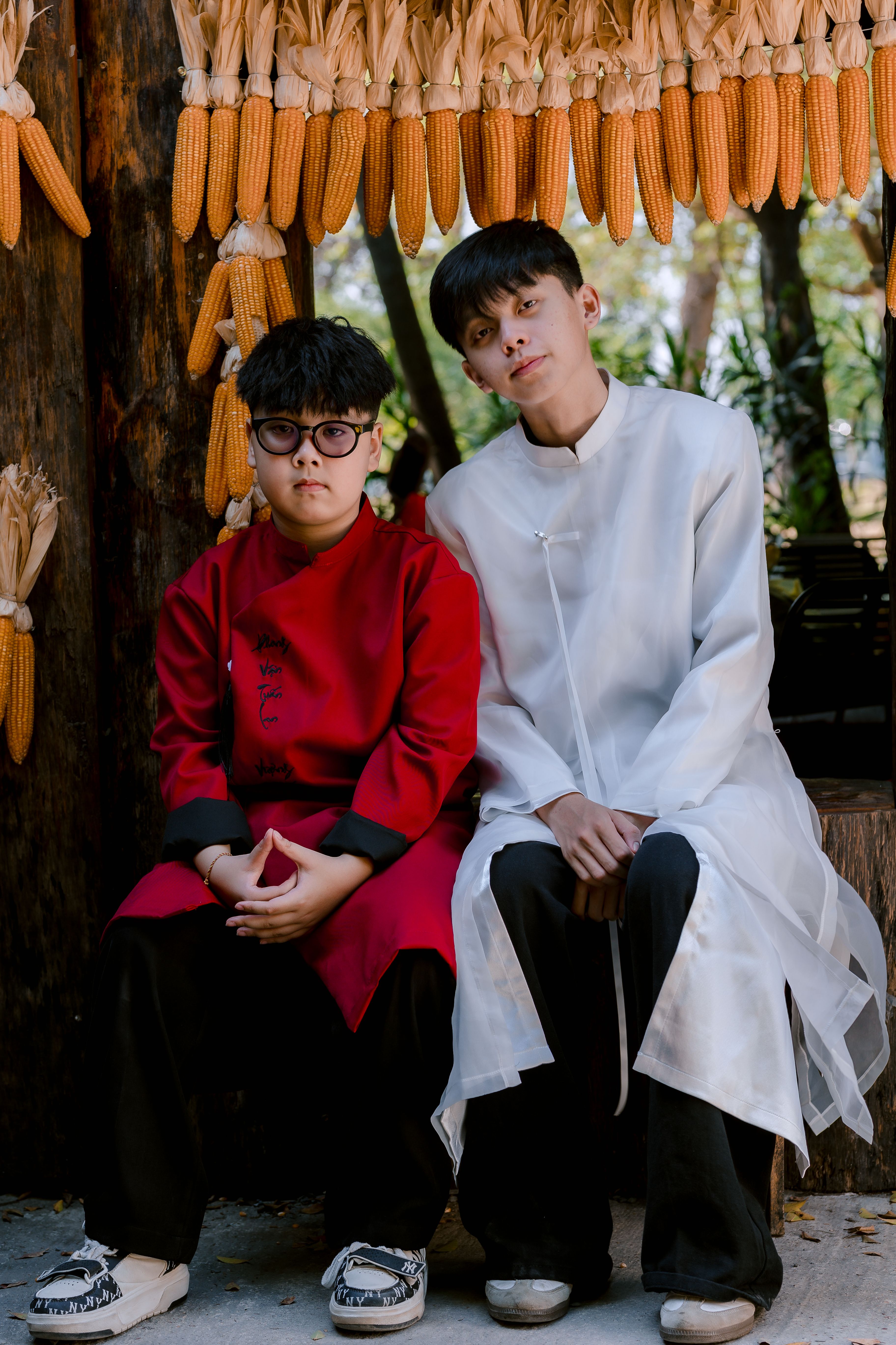 Two people in traditional Asian clothing posing near hanging corn decor outdoors.