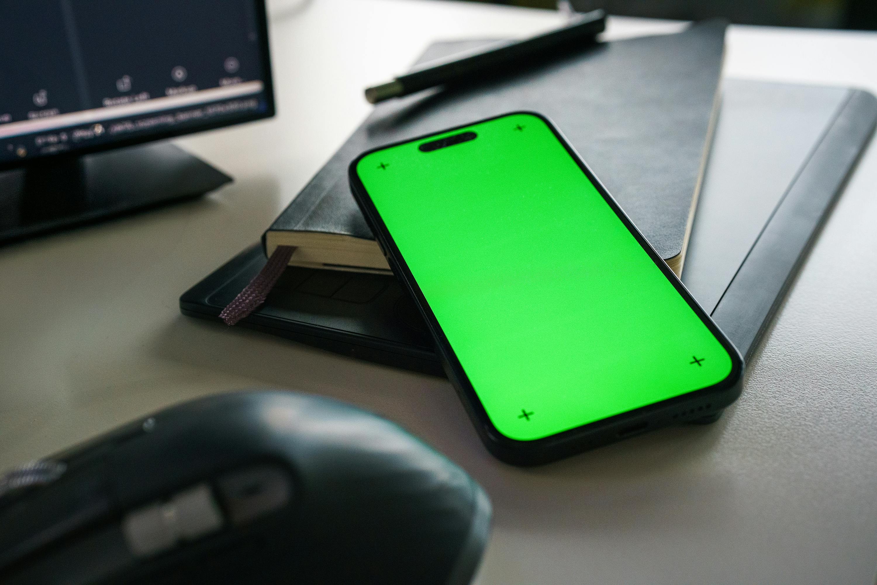 A clean and modern office desk with a smartphone displaying a green screen, notebook, and computer accessories.