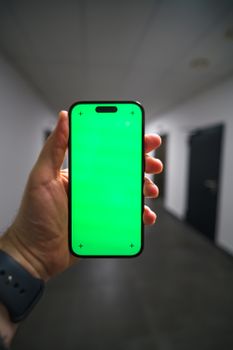 Hand holding a smartphone with a green screen in an office hallway, ideal for mockups.