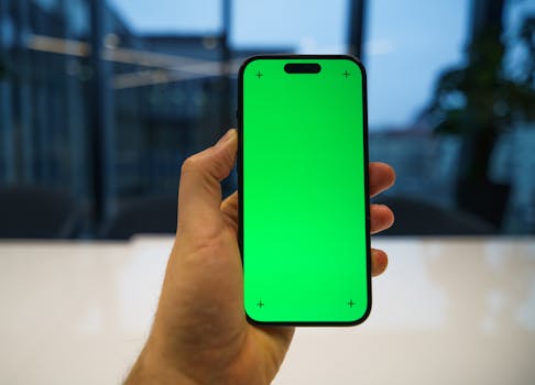 Hand holding smartphone with green chroma screen in office setting, ideal for mockup purposes.