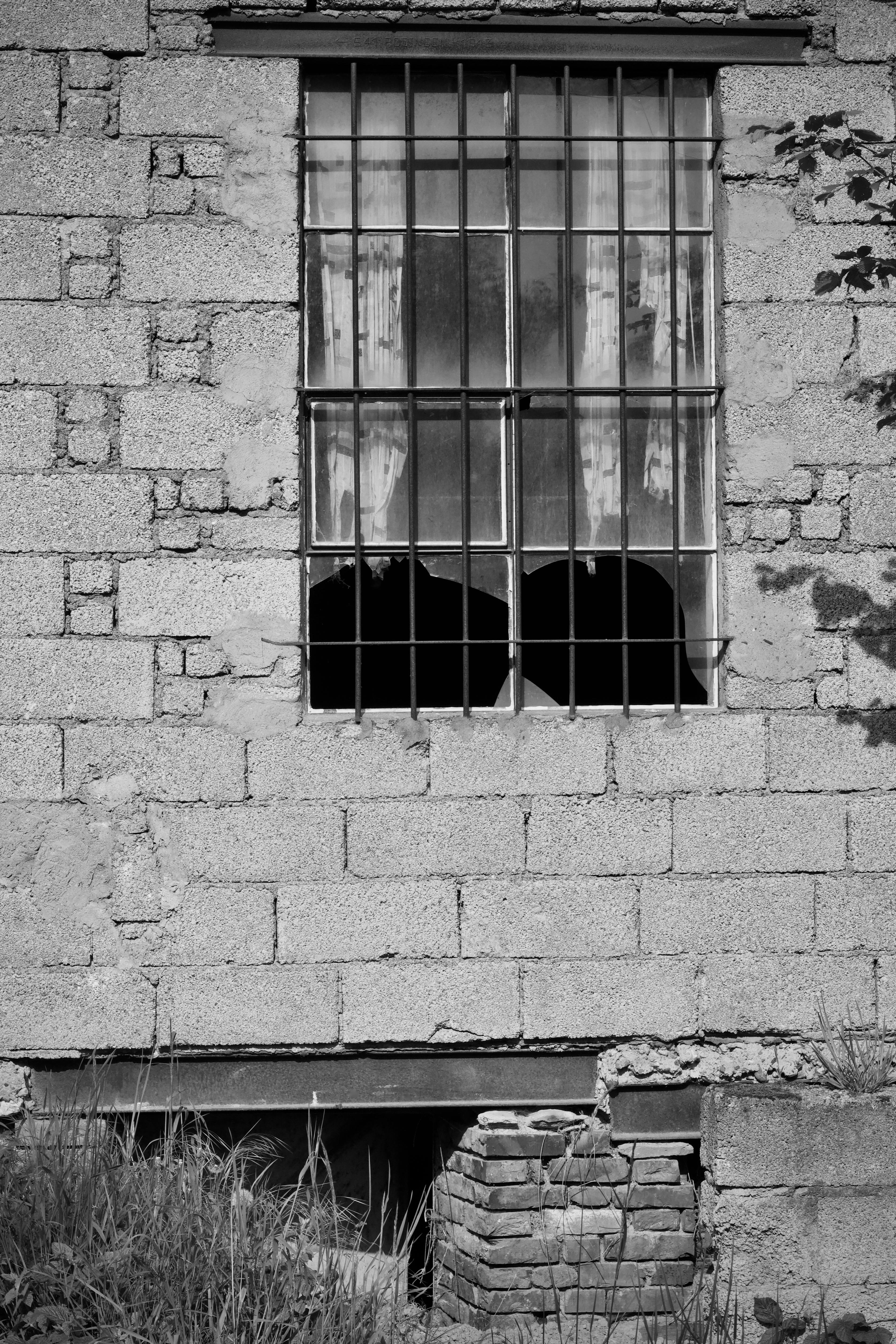 Gratis Immagine monocromatica di una finestra sbarrata con vetri rotti in un muro di cemento, a simboleggiare l'abbandono. Foto a disposizione