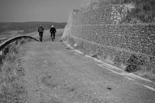 Two cyclists riding along a rural road lined with stone walls in a black and white setting.