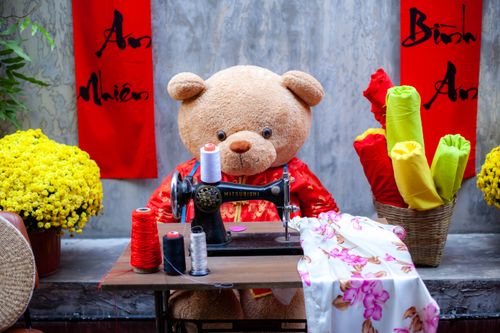 Free Charming teddy bear sewing with vintage machine surrounded by colorful fabric and flowers. Stock Photo