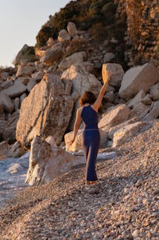 Woman in blue dress raising arm on rocky beach at sunset.