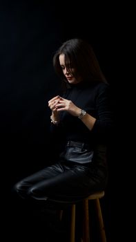 Elegant woman in black leather clothing sits contemplatively against a dark backdrop.