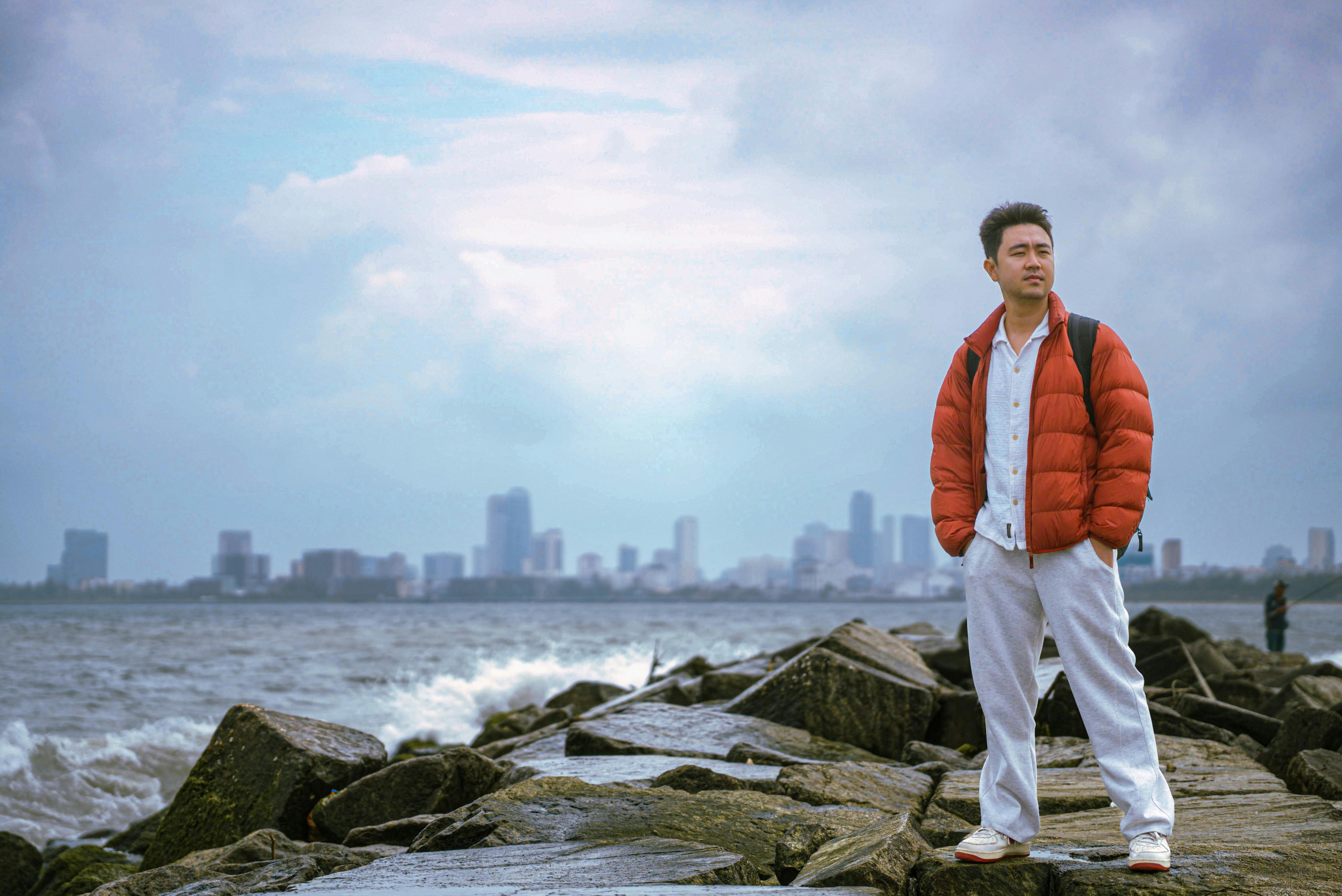 Gratis Un uomo con la giacca rossa in piedi sulle rocce vicino a Da Nang, con lo skyline sullo sfondo, mostra un paesaggio urbano costiero. Foto a disposizione