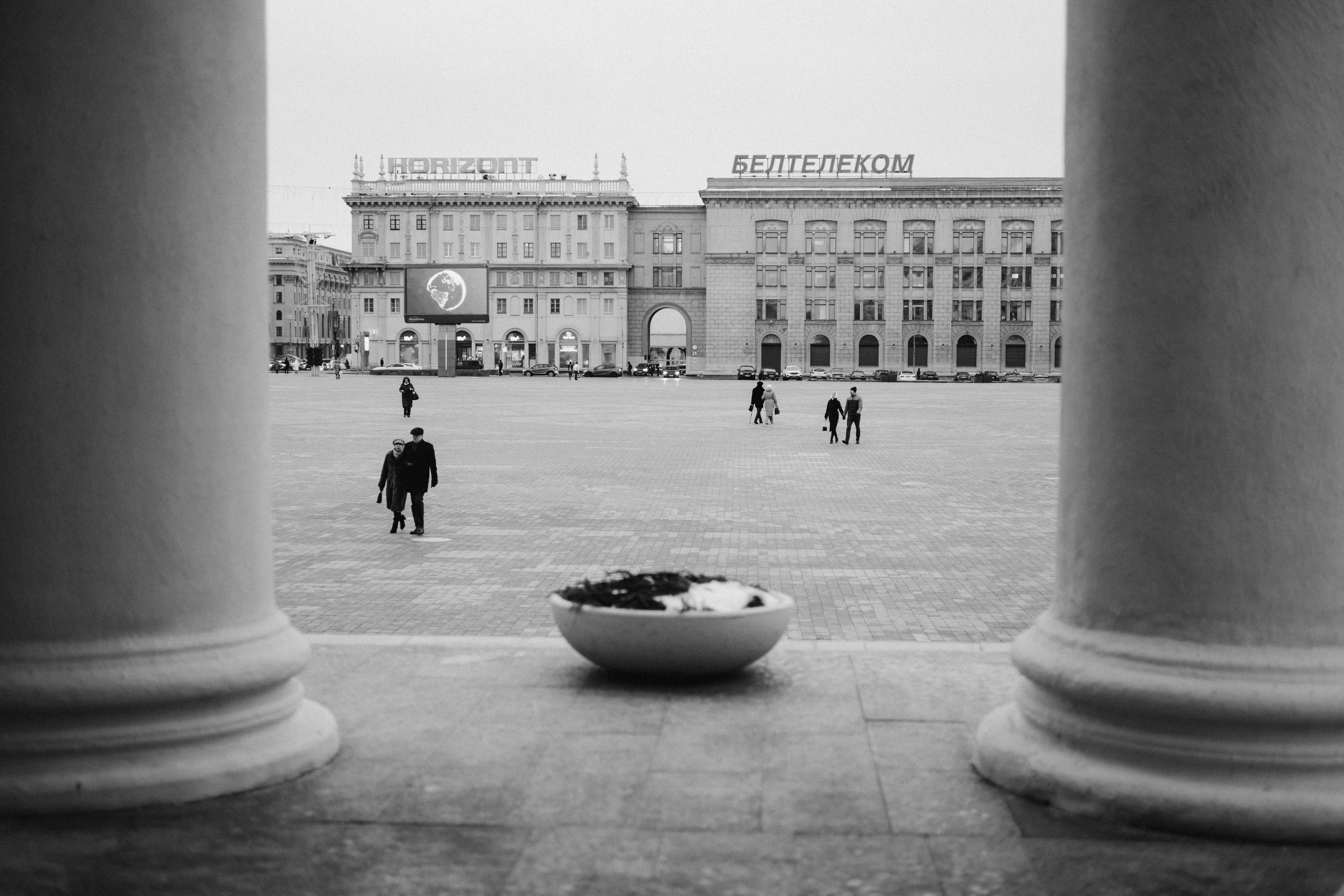 ฟรี ภาพถ่ายขาวดำของจัตุรัสในเมืองที่ล้อมรอบด้วยเสา แสดงให้เห็นผู้คนกำลังเดินอยู่ คลังภาพถ่าย