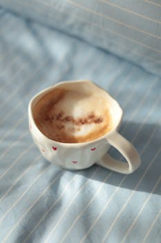 Top view of a white coffee cup with frothy cappuccino on blue striped bed linen, perfect for cozy mornings.