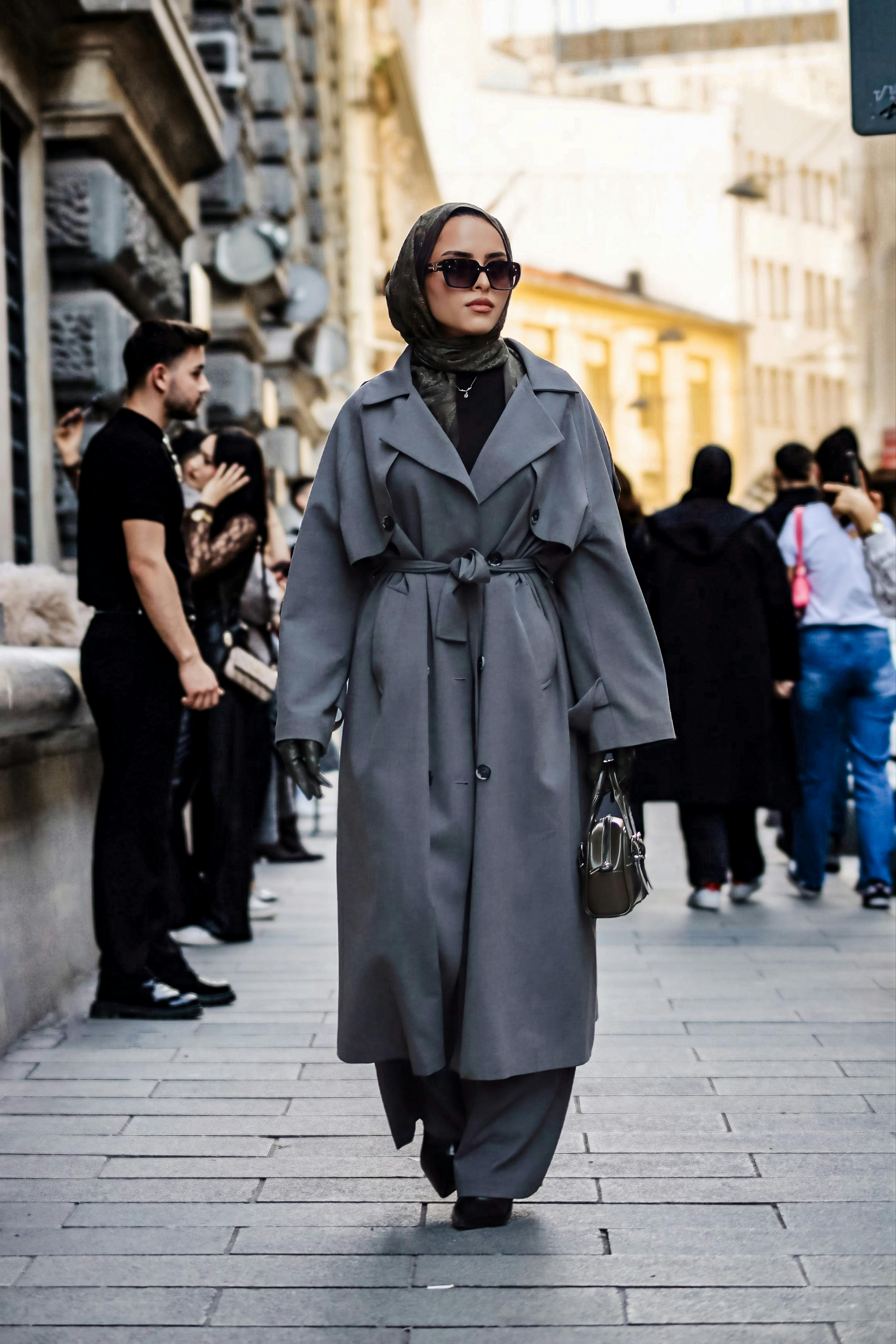 ฟรี หญิงสาวสง่างามในเสื้อโค้ทสีเทาและฮิญาบกำลังเดินอยู่บนถนนในเมืองที่พลุกพล่าน คลังภาพถ่าย