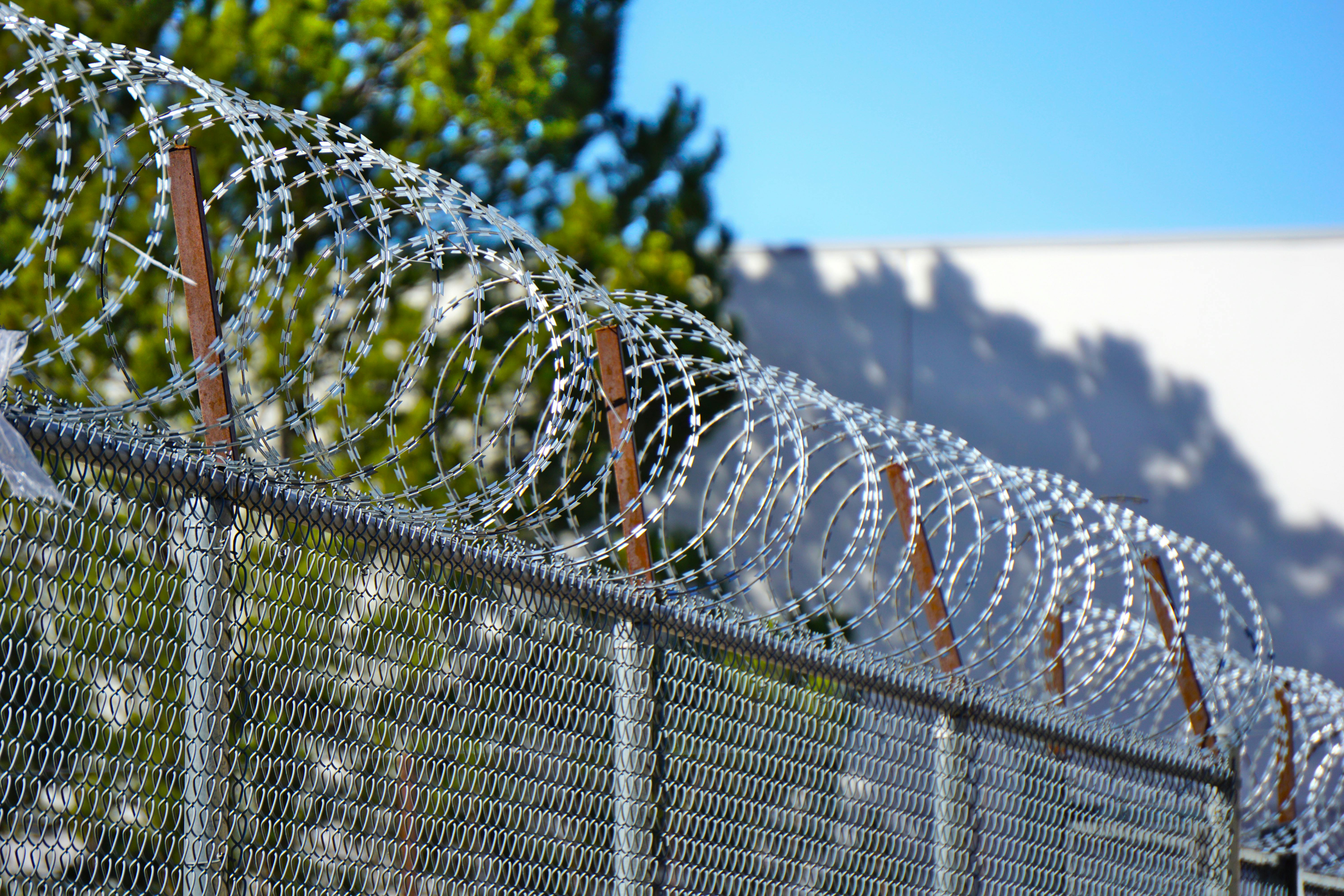 Razor wire coils run along the top of a chain-link security fence against a clear blue sky