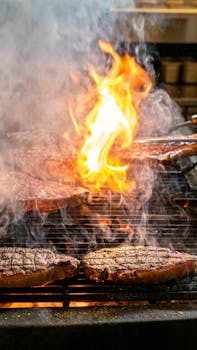 Sizzling steaks cooking on an open flame barbecue with smoke and fire.